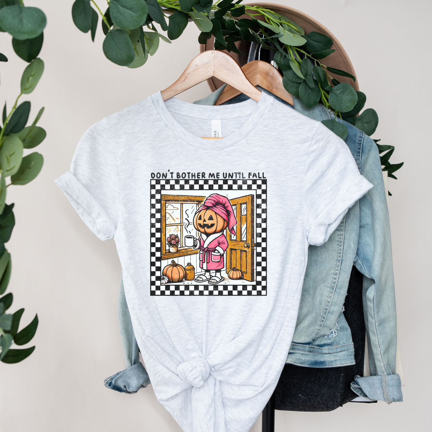 t-shirt featuring a cozy pumpkin character wearing a pink robe and towel, holding a coffee mug with the phrase "Don't Bother Me Until Fall," surrounded by autumn-inspired details and a checkered border. don't bother me until fall shirt, halloween pumpkin shirt, cozy autumn tee, fall lover t-shirt, pumpkin spice season shirt, spooky season apparel, cute pumpkin graphic tee, halloween aesthetic clothing, autumn lover gift, fall festival shirt, pumpkin patch outfit, cozy season clothing, spooky cute graphic te