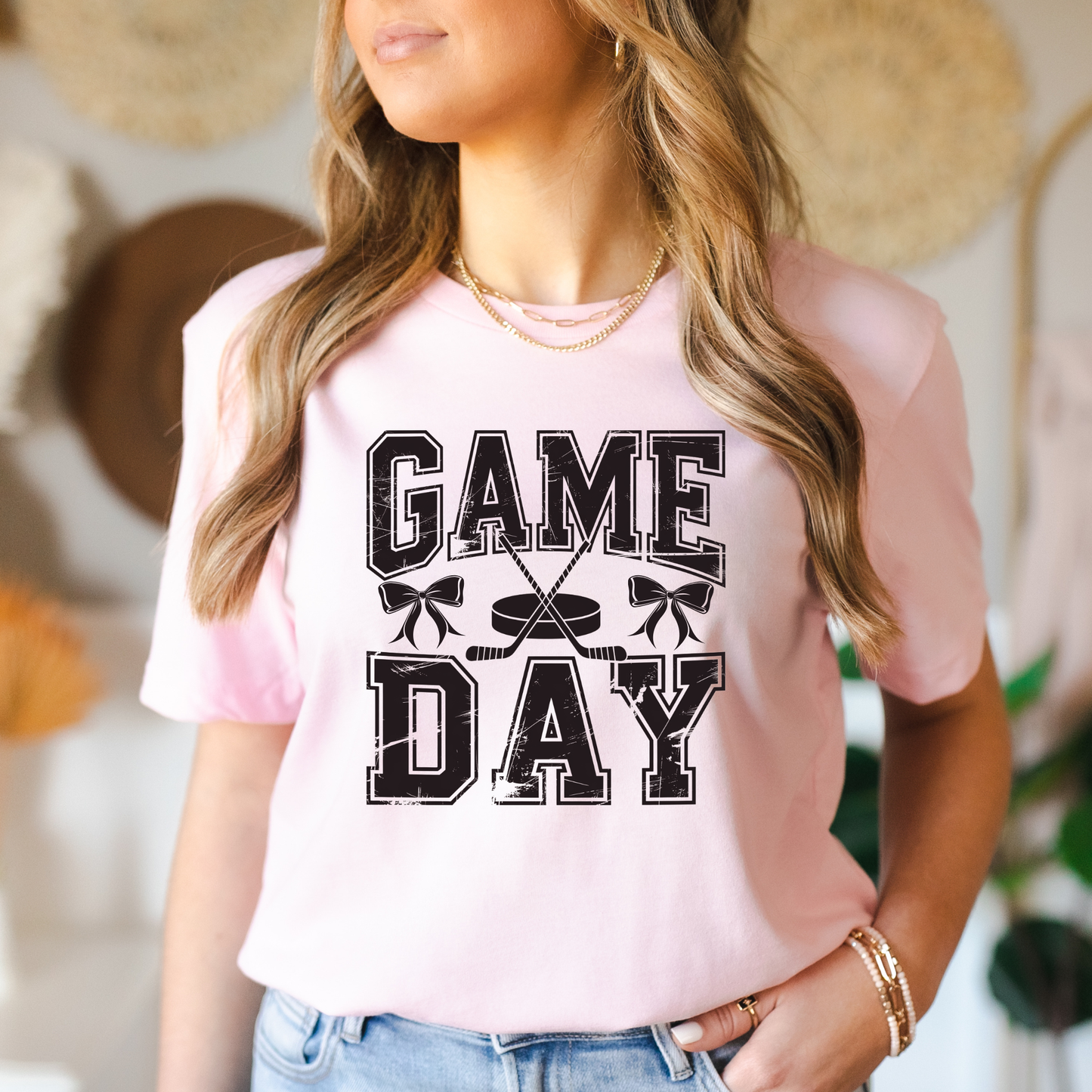 A woman wearing a light pink short-sleeve t-shirt with a distressed black graphic that reads 'GAME DAY' with crossed hockey sticks and hockey masks. She is wearing blue jeans and gold jewelry, with a softly blurred indoor background featuring boho decor.