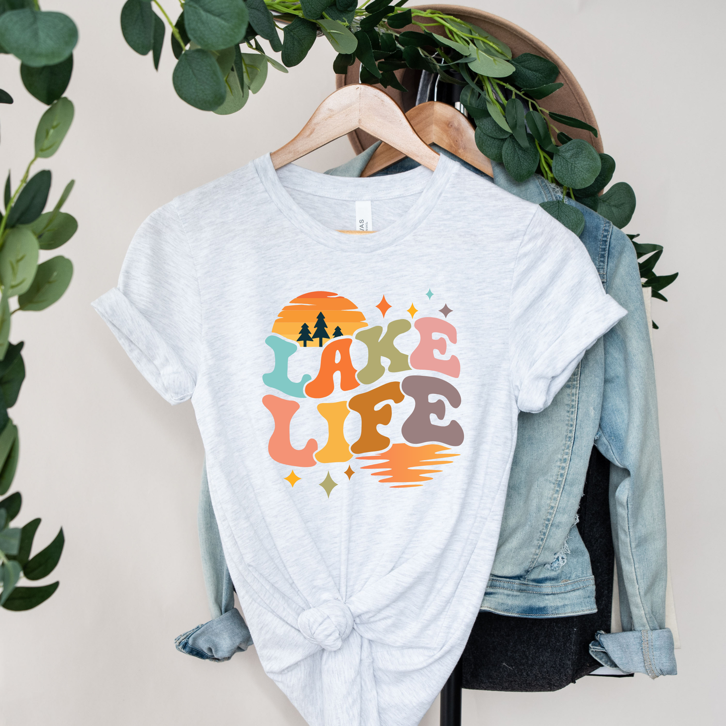 White t-shirt with 'Lake Life' design on a hanger with a denim jacket and greenery. lake life shirt, lake life tee, retro lake graphic shirt, sunset lake shirt, boating shirt, lake lover apparel, summer lake tee, unisex lake life clothing, lake house gift, fishing shirt, cottage lake outfit, lake vacation tee, boater shirt, lakeside apparel, retro sunset graphic tee