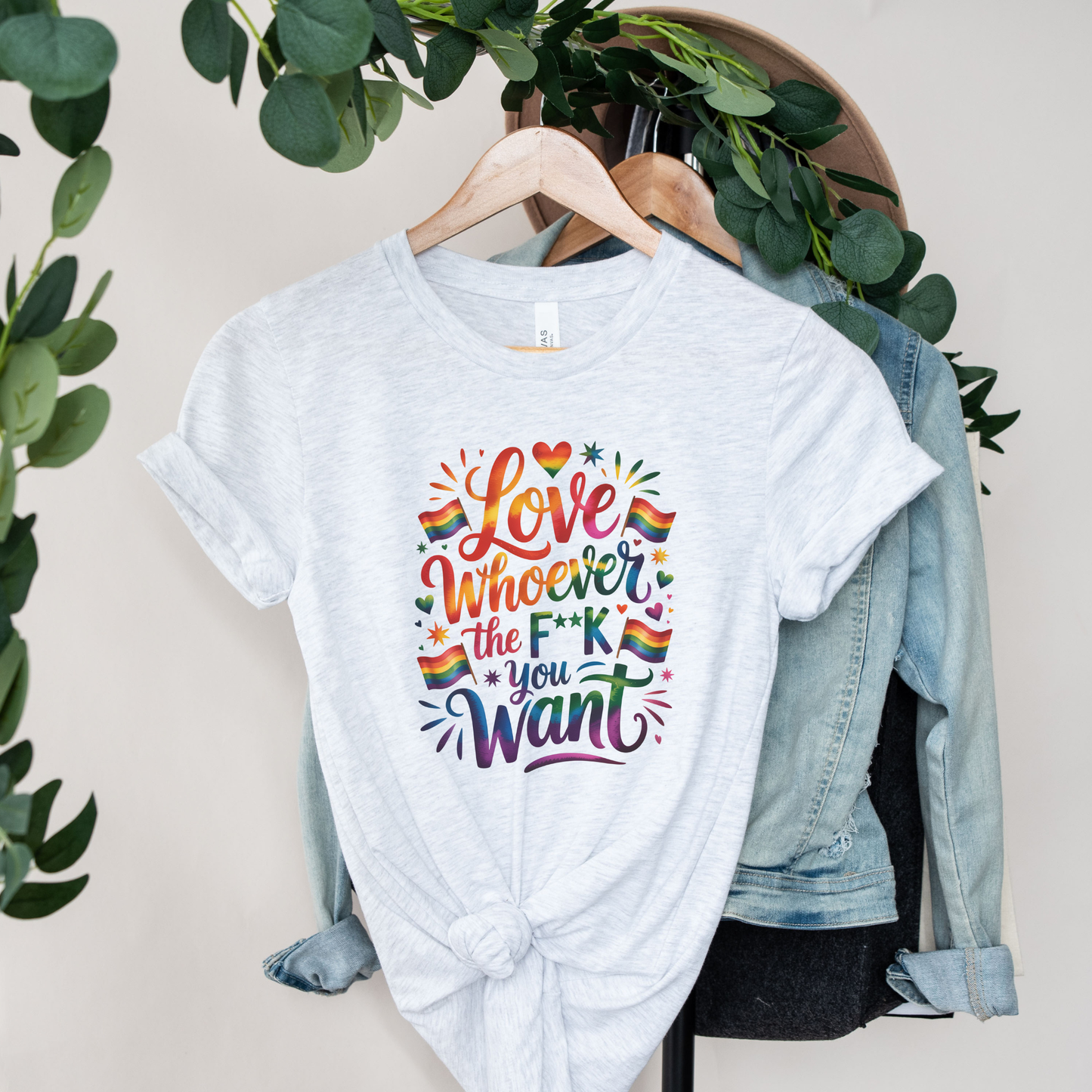 White t-shirt with colorful text and rainbow flags on a hanger with a denim jacket and greenery in the background. love is love, pride shirt, equality apparel, inclusive clothing, feminist tee, graphic shirt, LGBTQ+ fashion, Indigenous brand, Toronto streetwear, statement tee, self-expression shirt