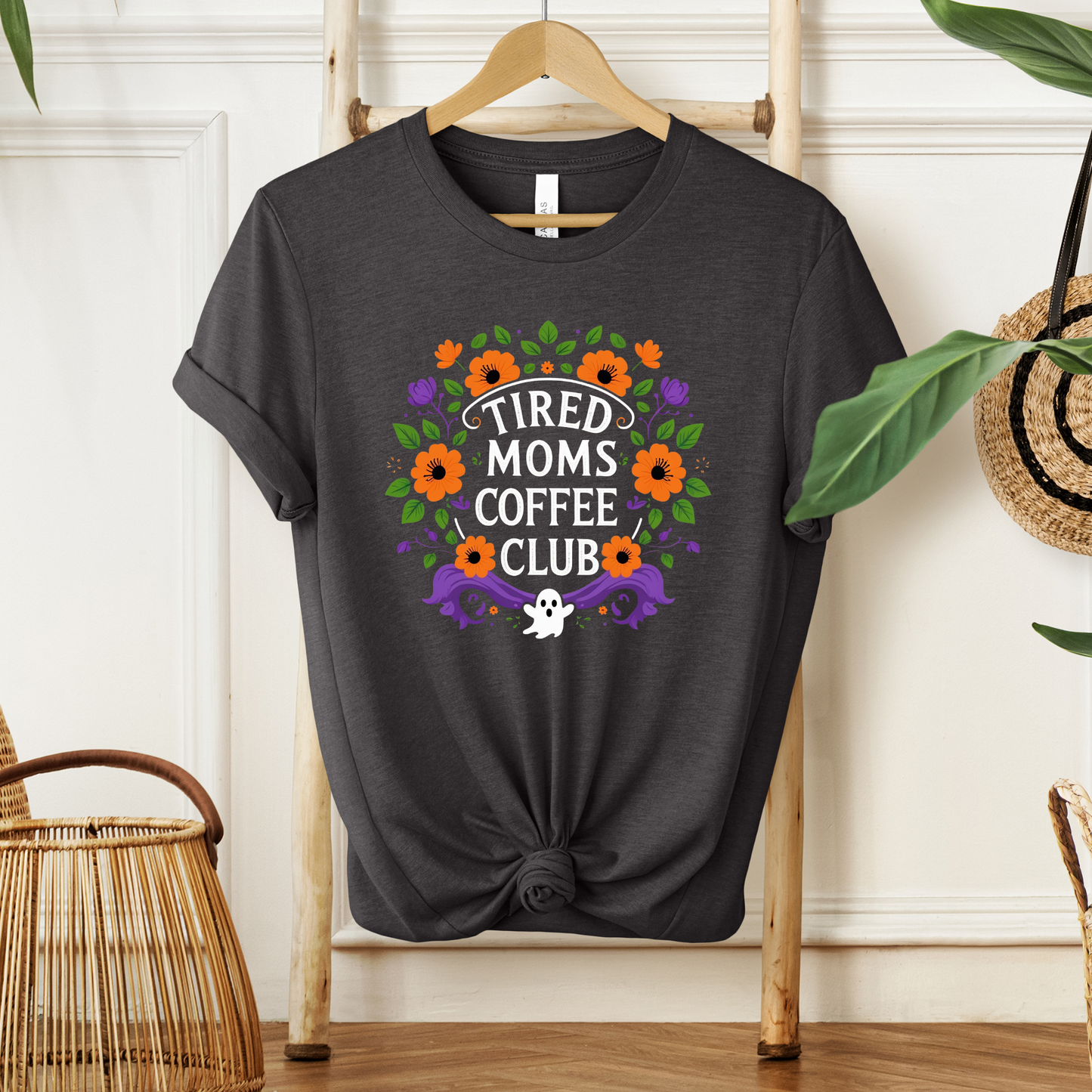 A dark charcoal gray graphic t-shirt hanging on a wooden hanger against a white paneled wall, styled with a knot at the bottom hem. The shirt features a colorful graphic with orange flowers, green leaves, purple accents, and a white ghost with the text 'TIRED MOMS COFFEE CLUB'.