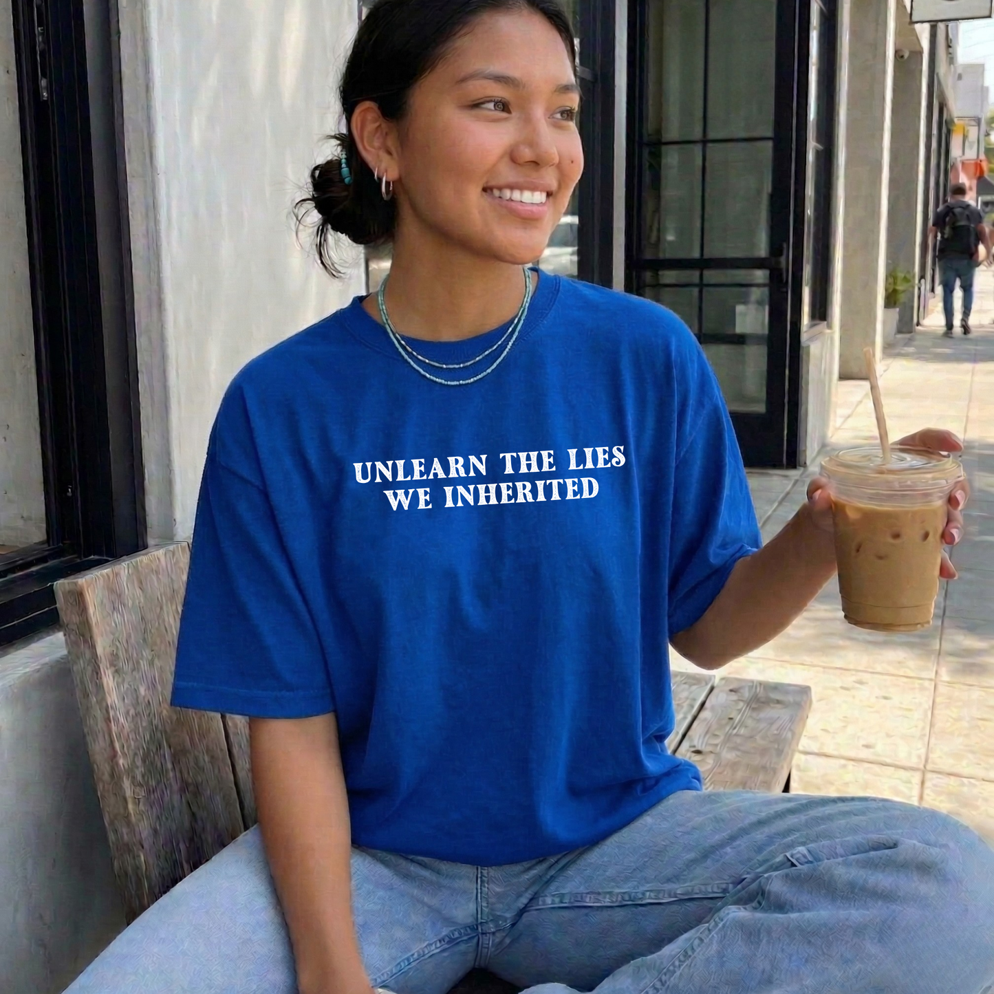 A woman sitting on a bench at a cafe wearing a bright blue t-shirt with the white printed text 'UNLEARN THE LIES WE INHERITED'. She is holding an iced coffee. unlearn the lies we inherited, truth and reconciliation shirt, indigenous shirt, social justice shirt, decolonize shirt, history matters shirt, educational shirt, activist shirt, statement tee, indigenous ally shirt, minimalist graphic tee, teacher shirt, advocacy apparel