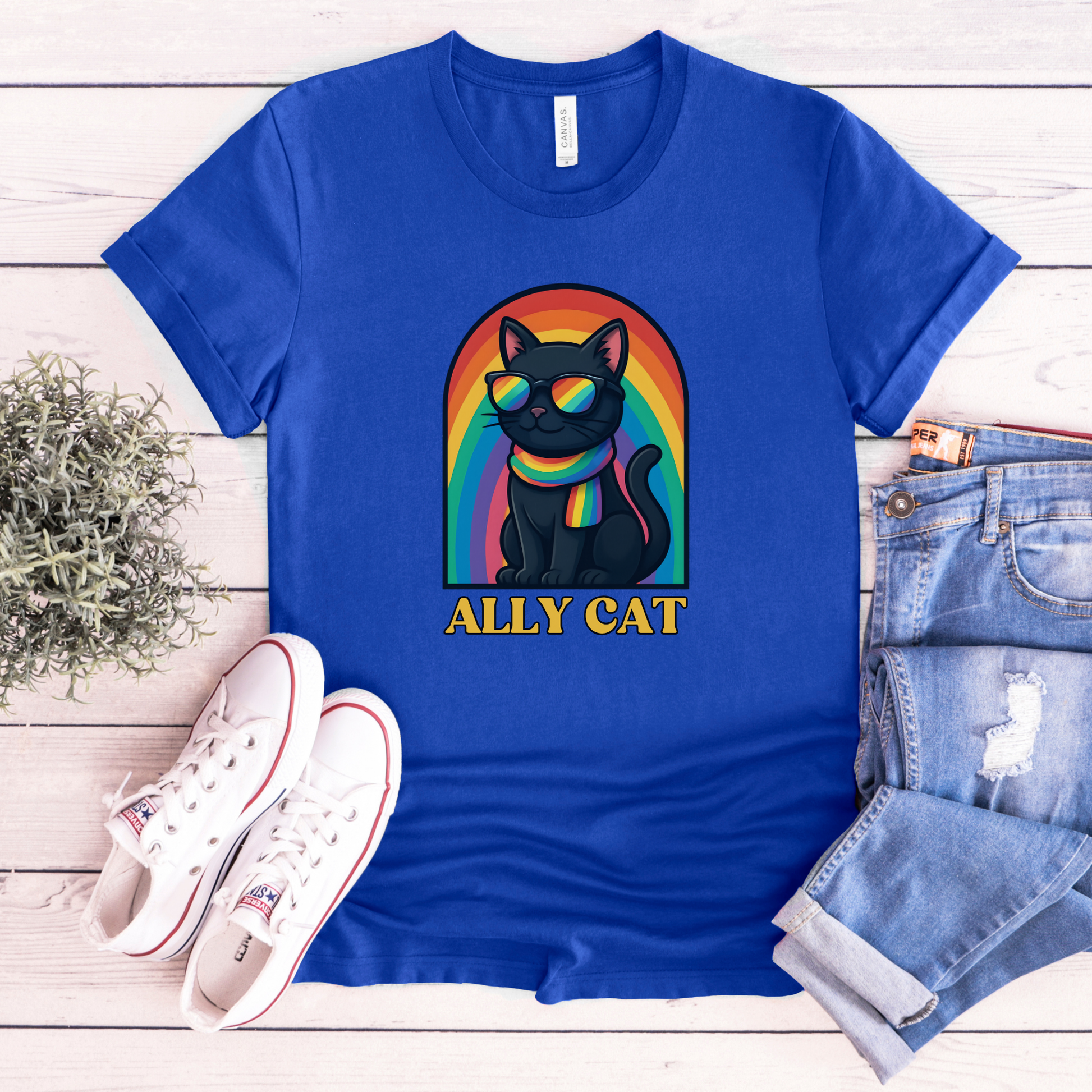 Blue t-shirt with 'ALLY CAT' graphic on a wooden surface with sneakers and jeans. ally cat shirt, rainbow cat shirt, LGBTQ ally shirt, pride cat tee, allyship shirt, cat lover shirt, unisex graphic tee, rainbow shirt, pride apparel, support LGBTQ, fun ally tee, colorful cat shirt, statement tee, cat graphic shirt, inclusive clothing
