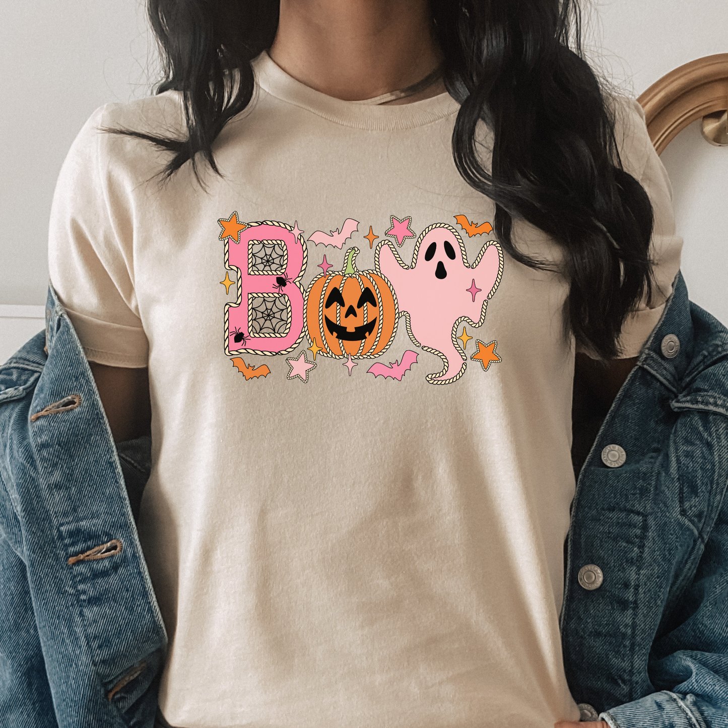 A person wearing a cream-colored t-shirt with a pink and orange Halloween graphic spelling 'BOO' with a pumpkin and ghost, styled with an open blue denim jacket. Boo Halloween Shirt, Cute Halloween Shirt, Pink Ghost Shirt, Pumpkin Halloween Tee, Spooky Season Shirt, Coquette Halloween Shirt, Fall Graphic Tee, Pumpkin Patch Outfit, Ghost Shirt, Halloween Aesthetic, Cute Fall Shirt, Women's Halloween Tee, Autumn Shirt.