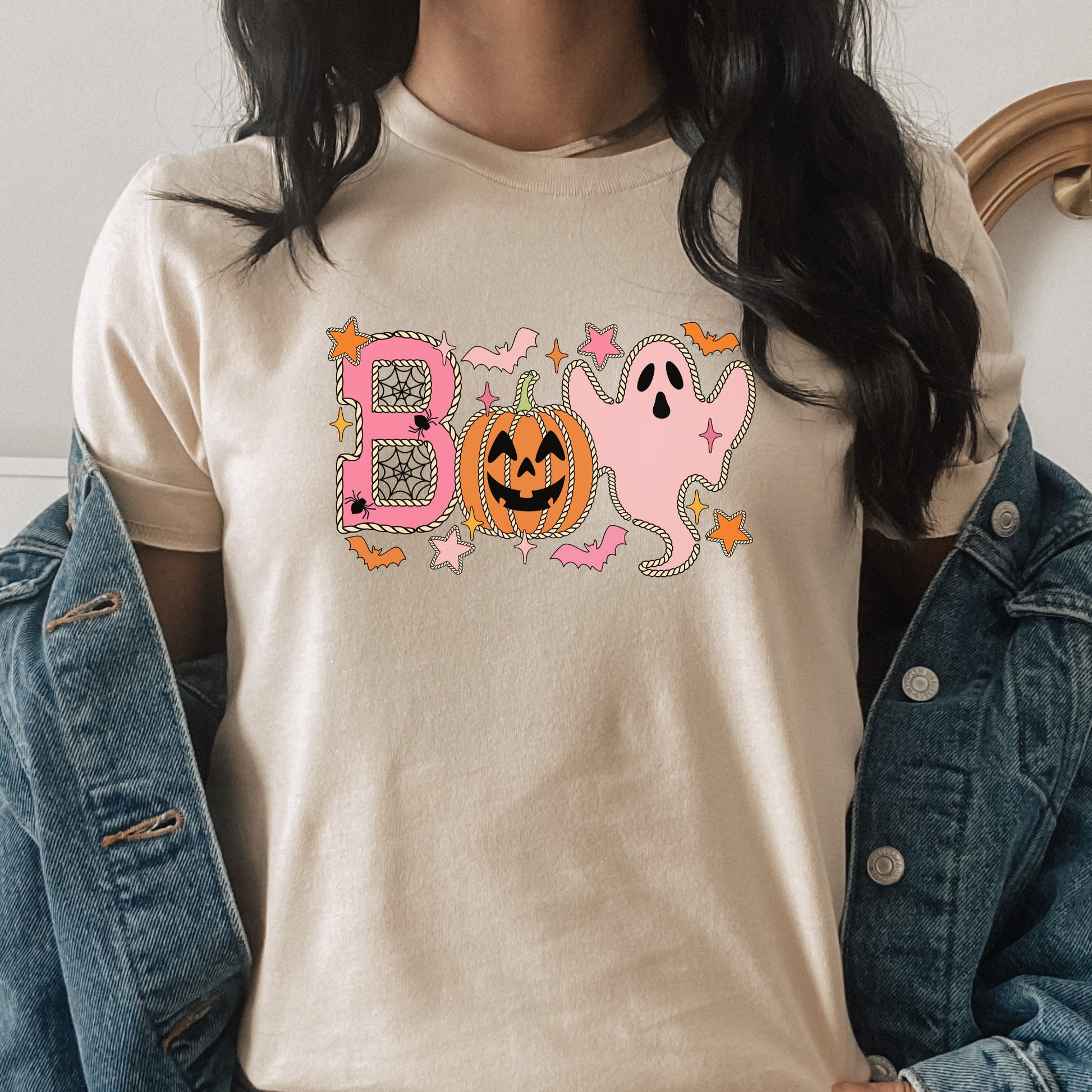 A person wearing a cream-colored t-shirt with a pink and orange Halloween graphic spelling 'BOO' with a pumpkin and ghost, styled with an open blue denim jacket. Boo Halloween Shirt, Cute Halloween Shirt, Pink Ghost Shirt, Pumpkin Halloween Tee, Spooky Season Shirt, Coquette Halloween Shirt, Fall Graphic Tee, Pumpkin Patch Outfit, Ghost Shirt, Halloween Aesthetic, Cute Fall Shirt, Women's Halloween Tee, Autumn Shirt.