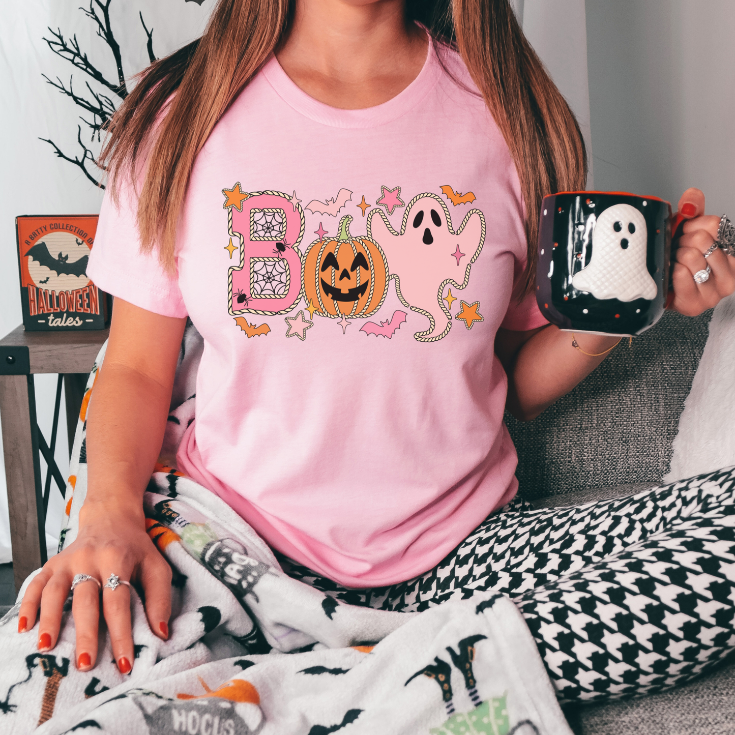 A woman wearing a light pink graphic t-shirt with a Halloween-themed 'Boo' design, sitting on a couch and holding a black mug with a white ghost. Boo Halloween Shirt, Cute Halloween Shirt, Pink Ghost Shirt, Pumpkin Halloween Tee, Spooky Season Shirt, Coquette Halloween Shirt, Fall Graphic Tee, Pumpkin Patch Outfit, Ghost Shirt, Halloween Aesthetic, Cute Fall Shirt, Women's Halloween Tee, Autumn Shirt.