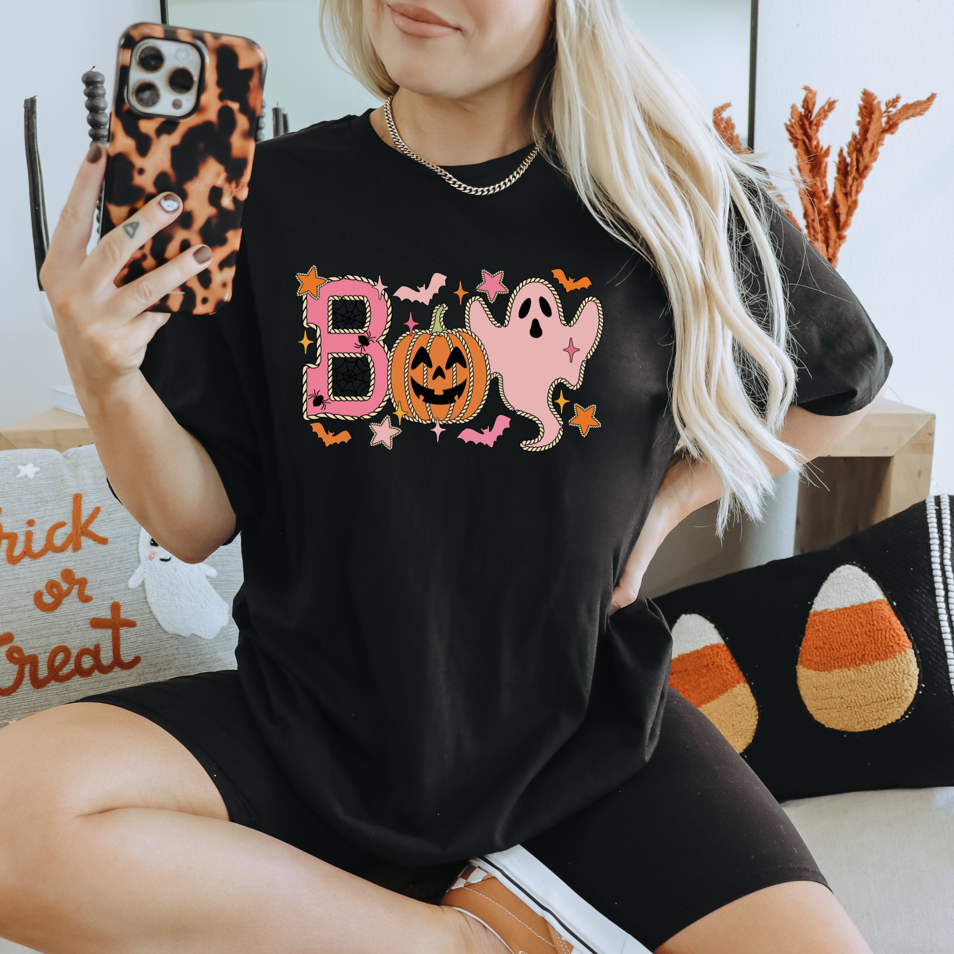A woman takes a mirror selfie wearing a black t-shirt with a colorful 'BOO' Halloween graphic featuring a jack-o'-lantern, ghost, and stars, paired with black biker shorts. Boo Halloween Shirt, Cute Halloween Shirt, Pink Ghost Shirt, Pumpkin Halloween Tee, Spooky Season Shirt, Coquette Halloween Shirt, Fall Graphic Tee, Pumpkin Patch Outfit, Ghost Shirt, Halloween Aesthetic, Cute Fall Shirt, Women's Halloween Tee, Autumn Shirt.