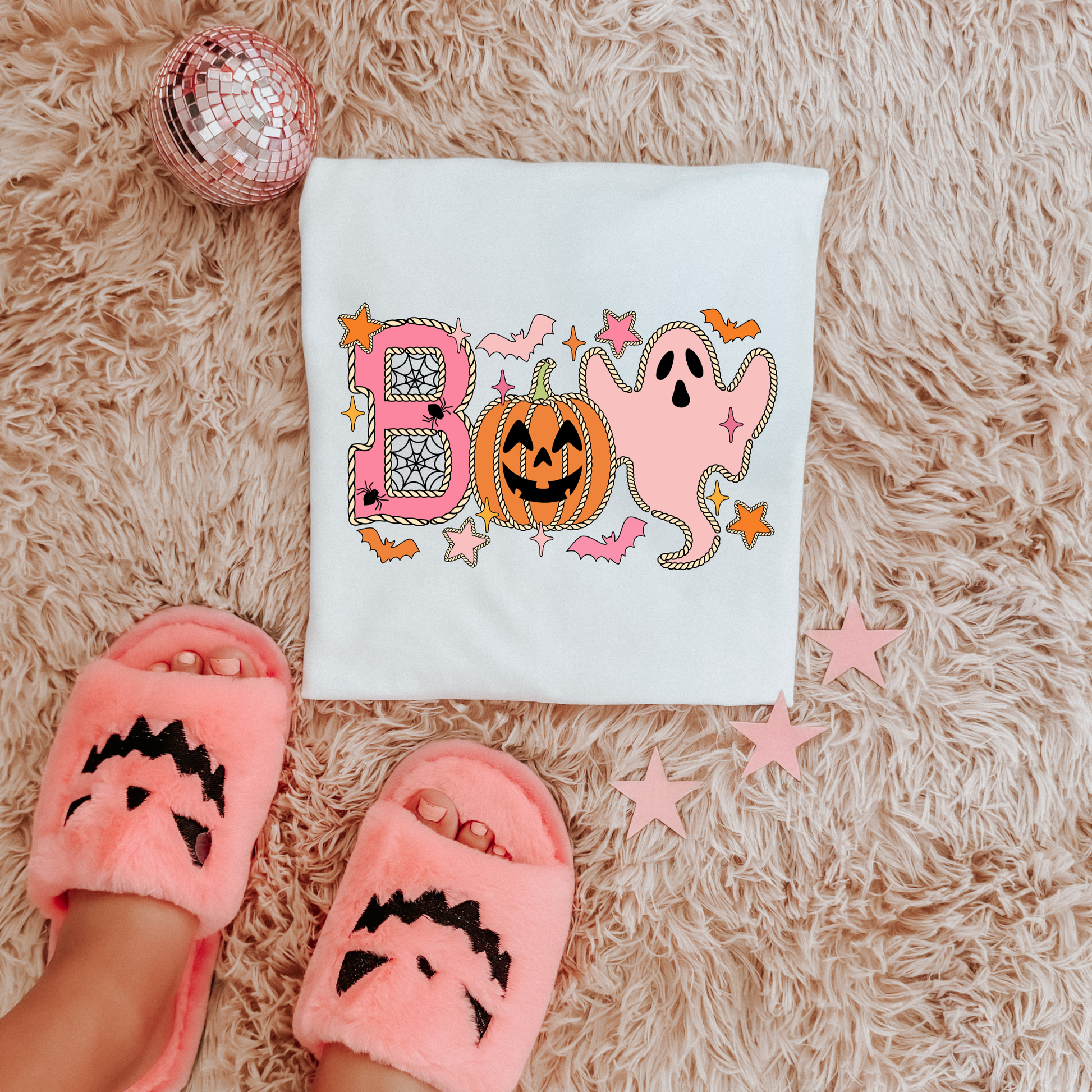 A white T-shirt laid flat on a fluffy cream rug, featuring a colorful Halloween graphic with the word 'BOO' in pink and orange, a jack-o'-lantern, and a ghost. A person is wearing pink fuzzy slide sandals with black face details, and decorative pink stars and a mirrored ball are placed around the shirt.