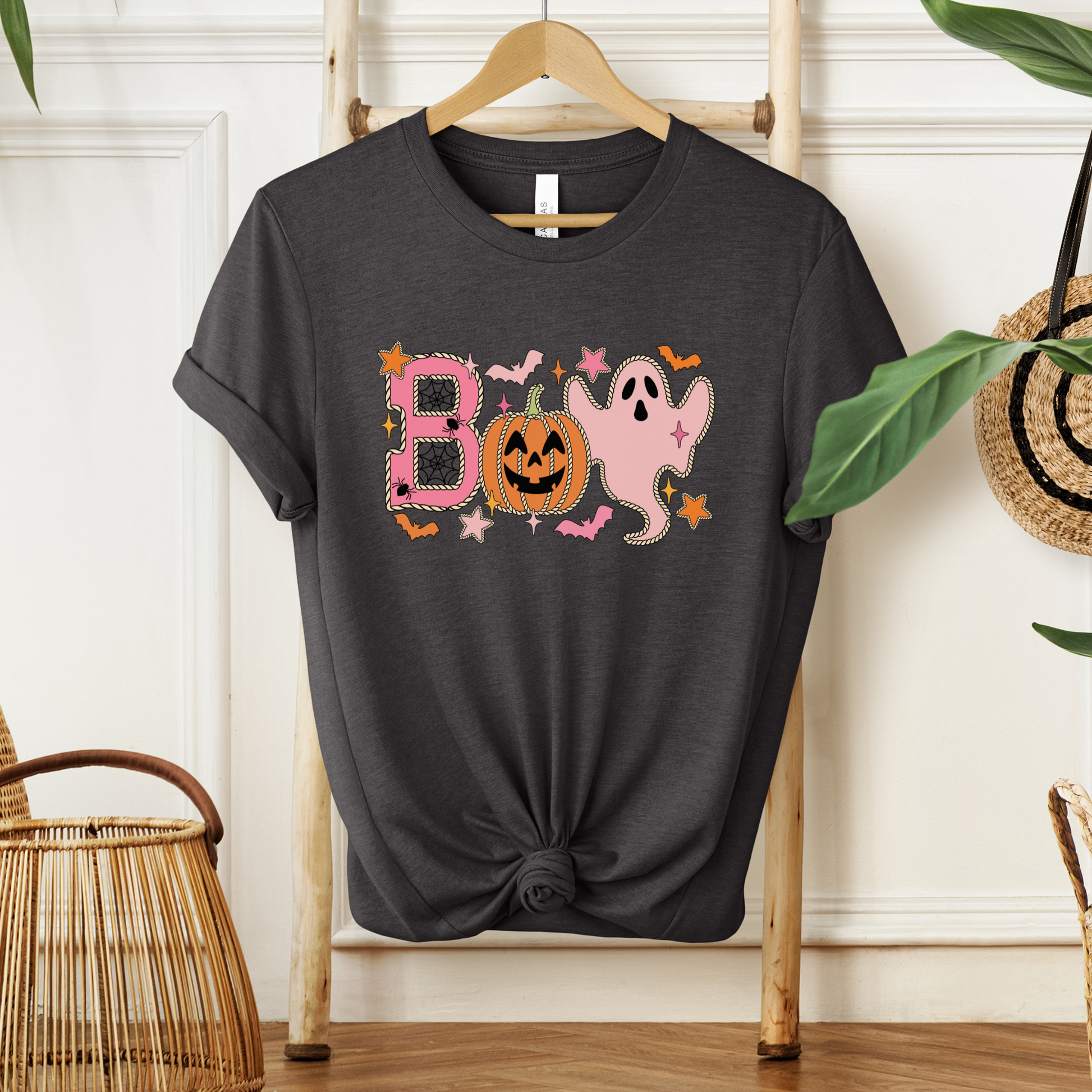 A dark charcoal gray graphic t-shirt with a knotted hem, hanging on a wooden hanger on a wooden ladder. The shirt features a colorful 'BOO' Halloween graphic with a pink ghost, orange jack-o'-lantern, and stars.