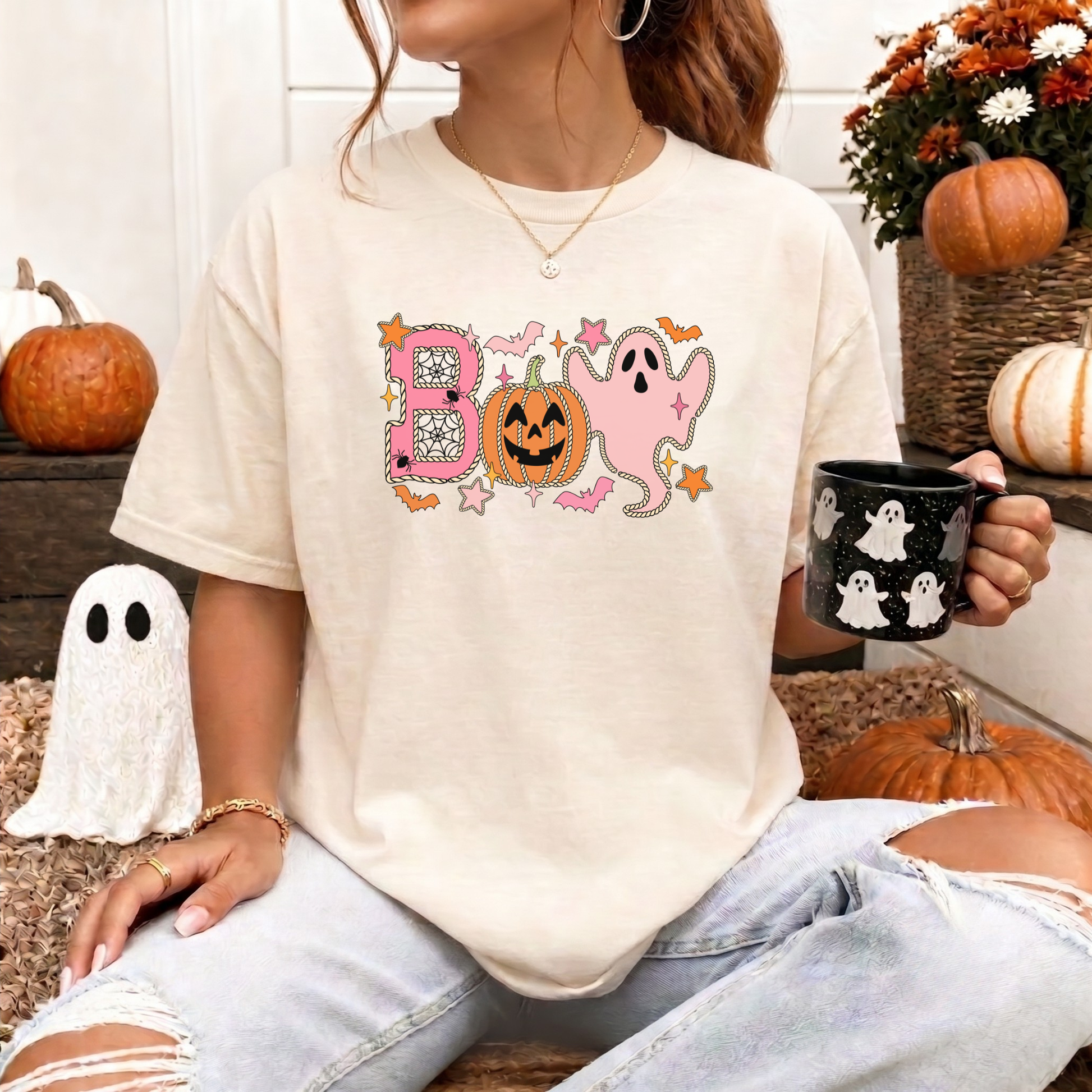 A woman wearing a cream-colored t-shirt with a colorful 'BOO' Halloween graphic, paired with ripped light-wash jeans, holding a black mug with white ghosts while sitting in a cozy autumn-themed setting with pumpkins. Boo Halloween Shirt, Cute Halloween Shirt, Pink Ghost Shirt, Pumpkin Halloween Tee, Spooky Season Shirt, Coquette Halloween Shirt, Fall Graphic Tee, Pumpkin Patch Outfit, Ghost Shirt, Halloween Aesthetic, Cute Fall Shirt, Women's Halloween Tee, Autumn Shirt.