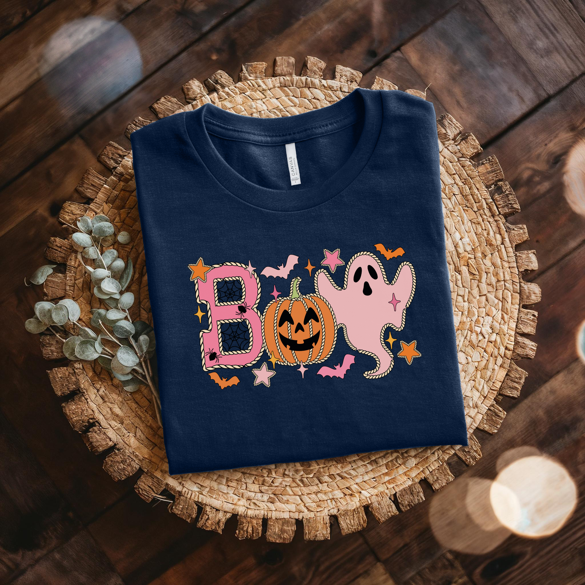 A navy blue folded t-shirt with a colorful Halloween-themed graphic spelling 'BOO' using a pumpkin, ghost, bats, and stars, styled on a woven placemat over a dark wood background.
