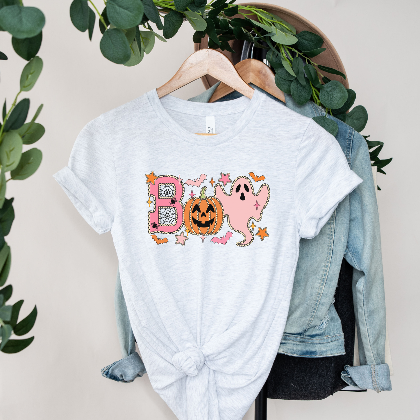 A light heather gray t-shirt with a 'BOO' Halloween graphic hanging on a wooden hanger, styled with a light blue denim jacket and dark clothing on a hanger stand, with green foliage hanging in the background.