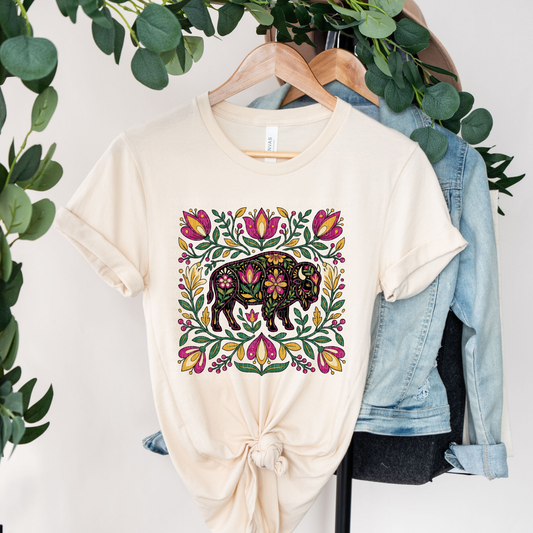A cream-colored t-shirt with a colorful floral bison graphic, styled on a wooden hanger with a denim jacket behind it and green foliage hanging around the top.
