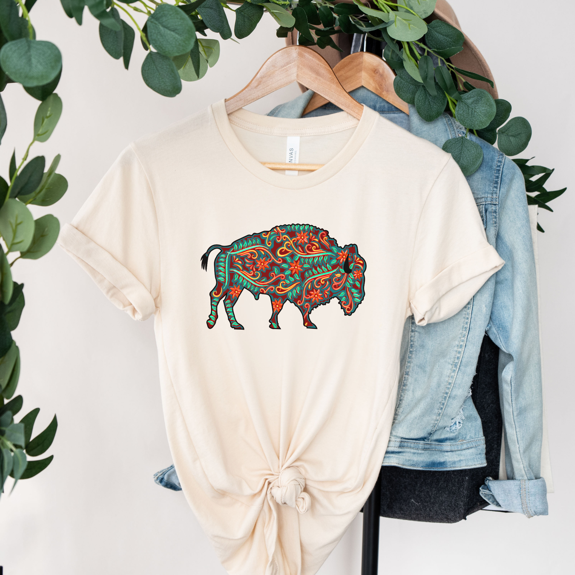 Beige t-shirt with a colorful buffalo design on a hanger with greenery and denim jeans in the background. native floral buffalo shirt, indigenous buffalo shirt, floral buffalo tee, indigenous design shirt, buffalo graphic tee, native inspired apparel, cultural pride shirt, unisex indigenous tee, floral indigenous shirt, buffalo and floral shirt, indigenous owned clothing, meaningful graphic tee