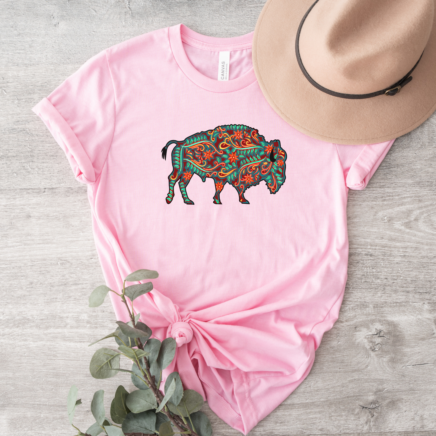 Pink t-shirt with a colorful buffalo design, accompanied by a hat and plant on a wooden surface. native floral buffalo shirt, indigenous buffalo shirt, floral buffalo tee, indigenous design shirt, buffalo graphic tee, native inspired apparel, cultural pride shirt, unisex indigenous tee, floral indigenous shirt, buffalo and floral shirt, indigenous owned clothing, meaningful graphic tee