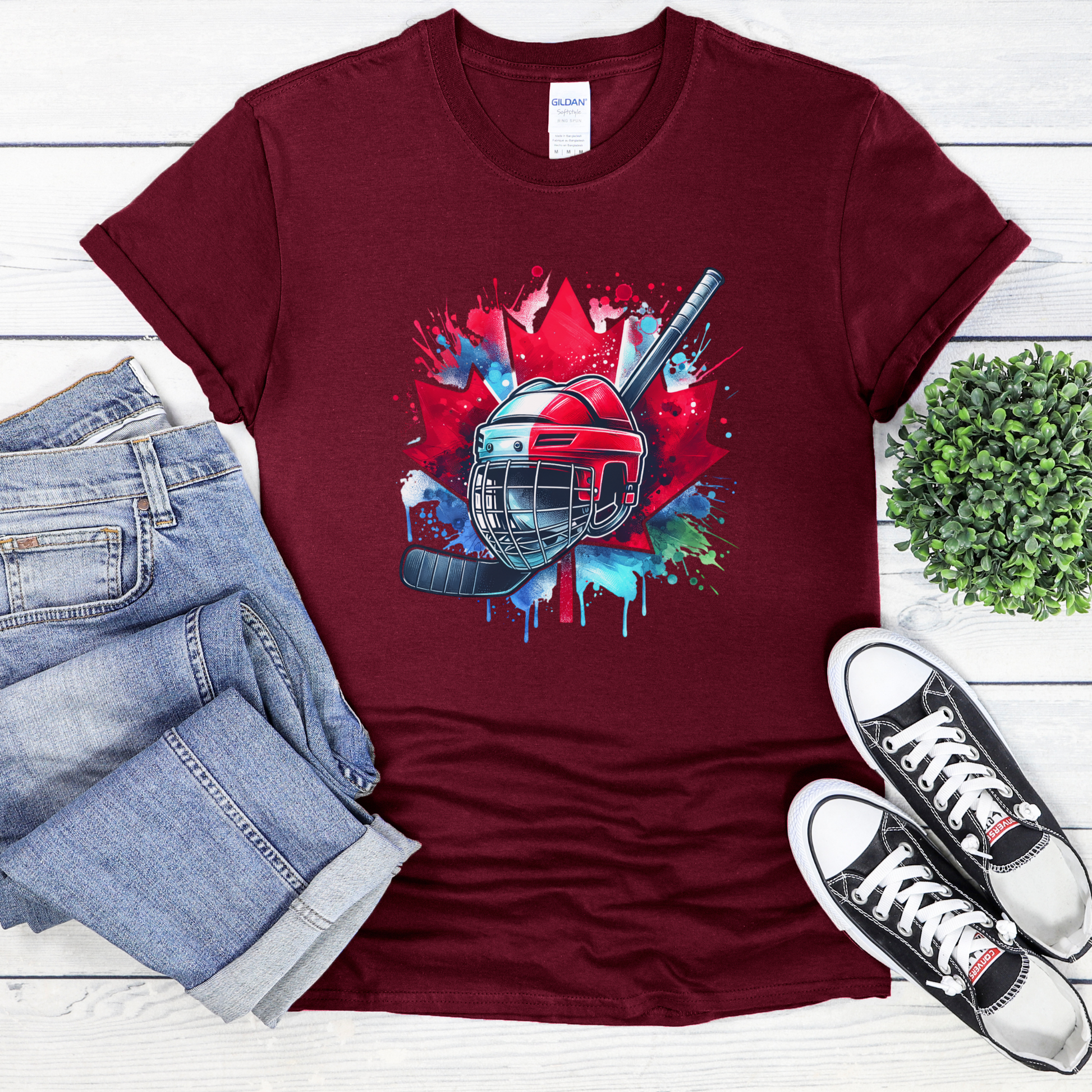 Canada Hockey Shirt featuring a colorful hockey helmet and stick graphic layered over a vibrant red Canadian maple leaf with paint splash accents. Proudly made in Canada, this patriotic hockey t-shirt is perfect for hockey fans, players, coaches, and Canada Day celebrations.