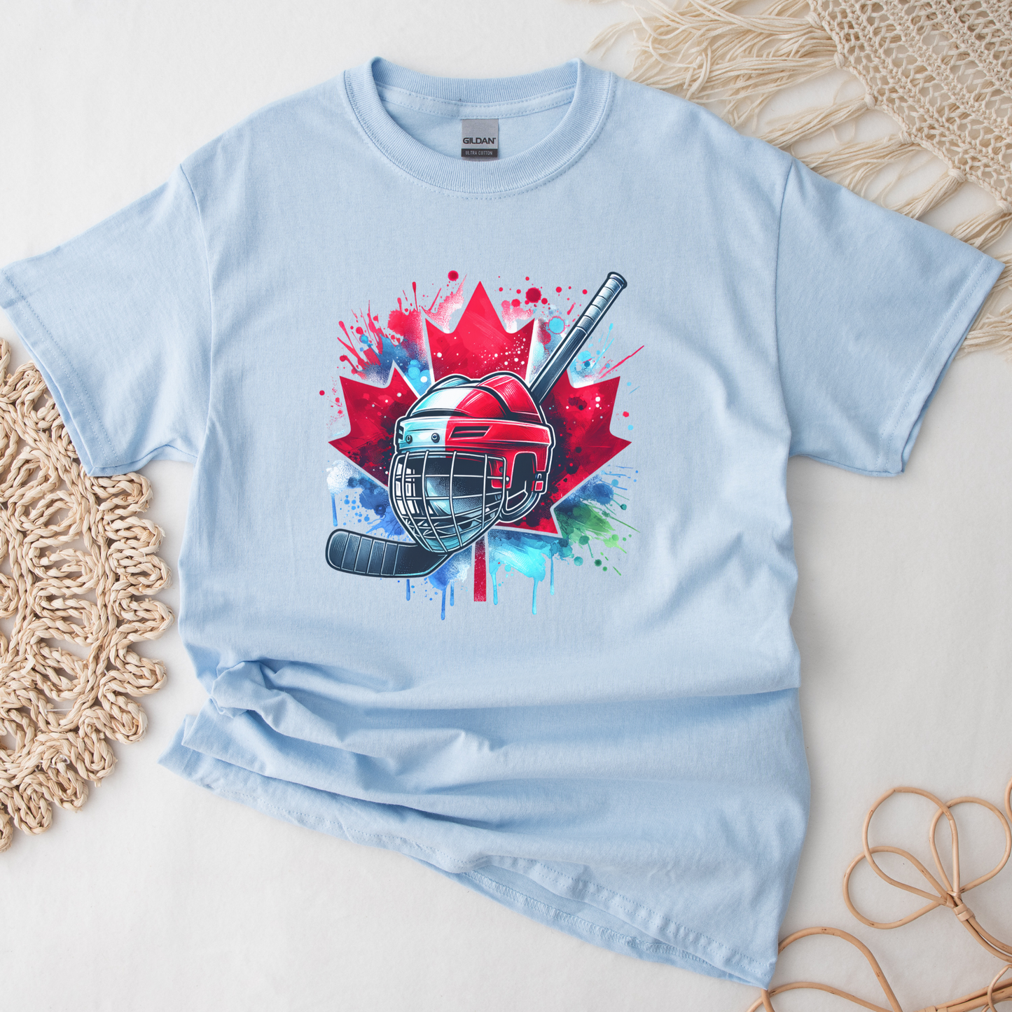 Canada Hockey Shirt featuring a colorful hockey helmet and stick graphic layered over a vibrant red Canadian maple leaf with paint splash accents. Proudly made in Canada, this patriotic hockey t-shirt is perfect for hockey fans, players, coaches, and Canada Day celebrations.
