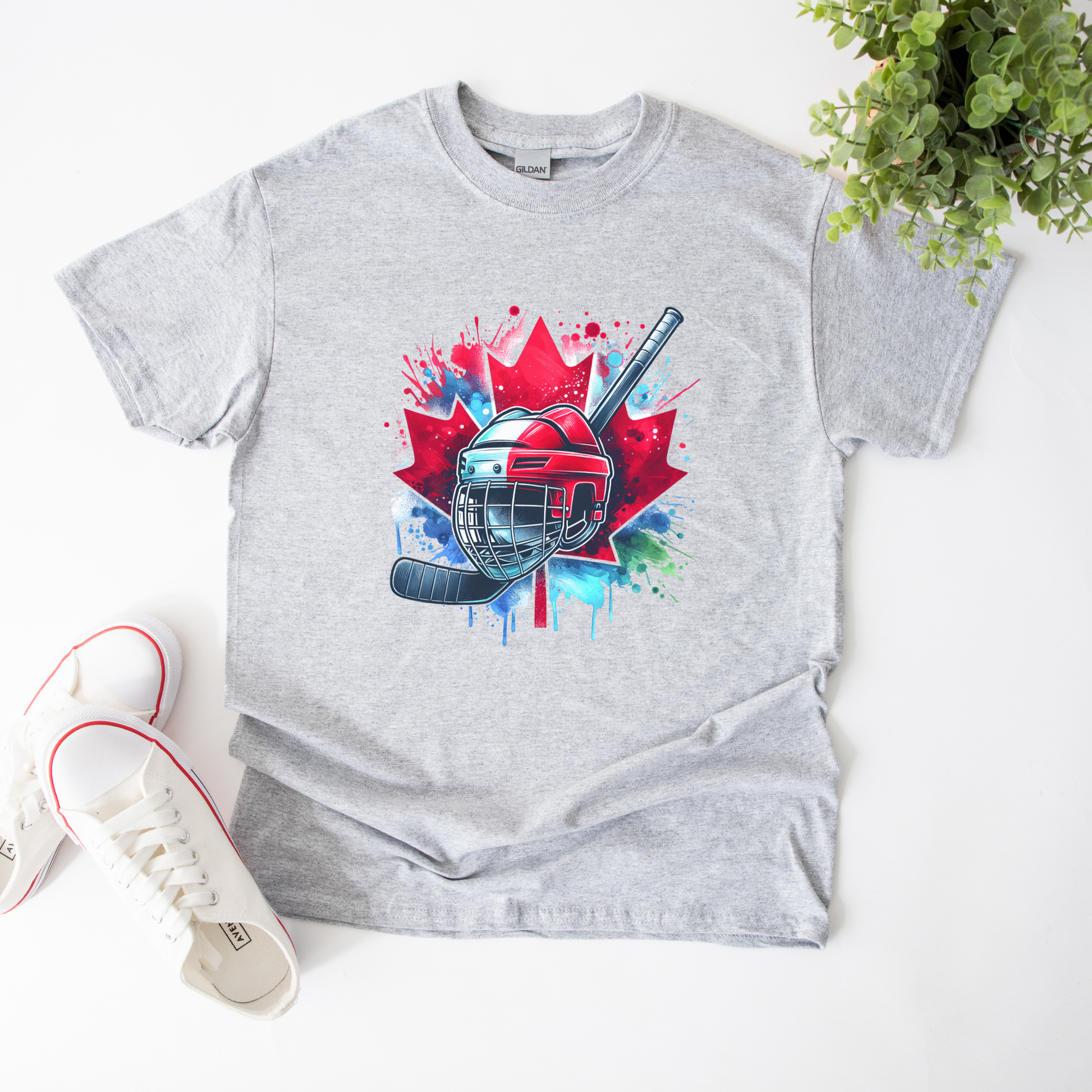 Canada Hockey Shirt featuring a colorful hockey helmet and stick graphic layered over a vibrant red Canadian maple leaf with paint splash accents. Proudly made in Canada, this patriotic hockey t-shirt is perfect for hockey fans, players, coaches, and Canada Day celebrations.