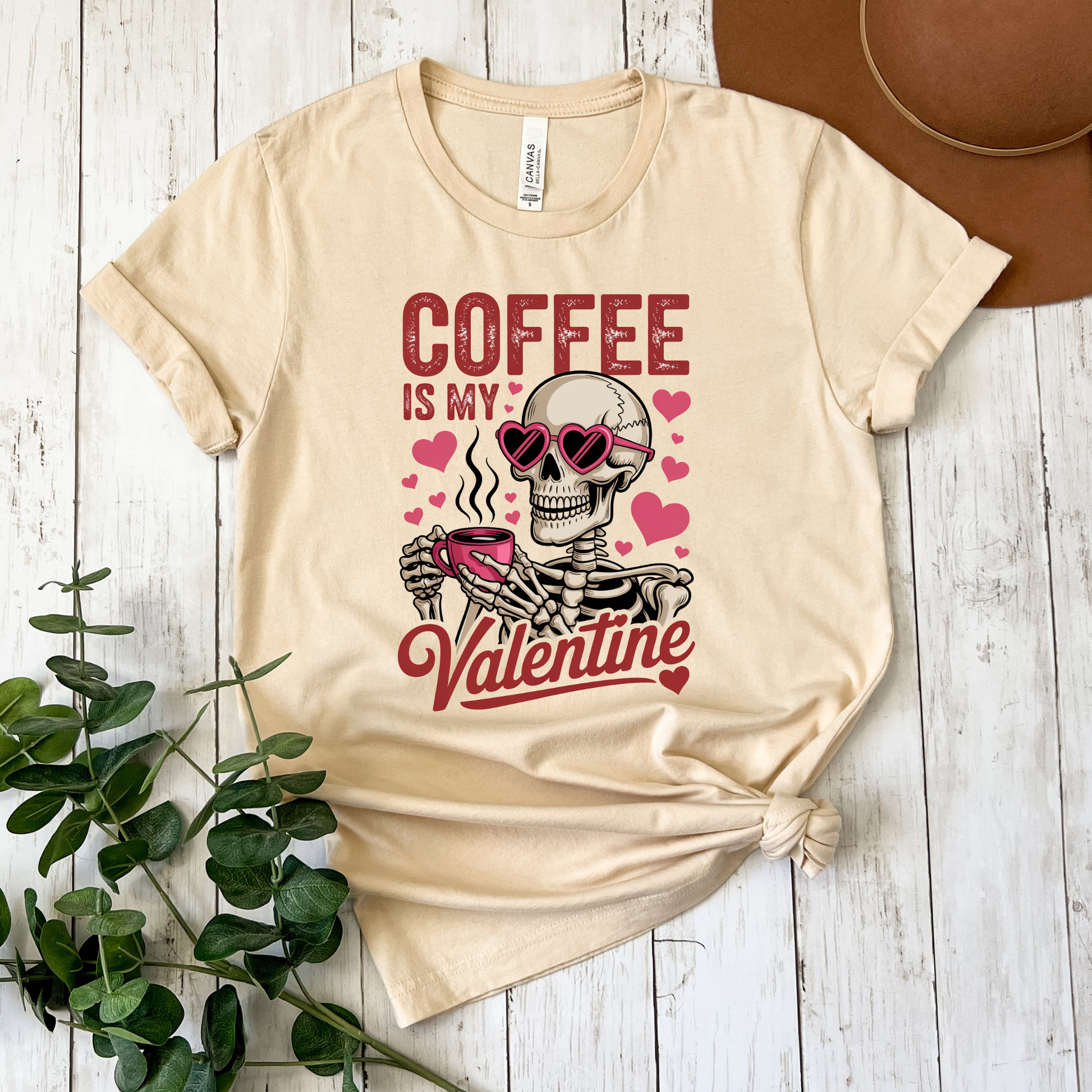 Beige t-shirt with skeleton holding a coffee cup and text 'Coffee is my Valentine' on a wooden surface. coffee shirt, coffee lover tee, valentine shirt, funny valentine shirt, coffee is my valentine, cute coffee shirt, unisex graphic tee, caffeine shirt, gift for coffee lover, funny t shirt, casual tee, valentines day shirt