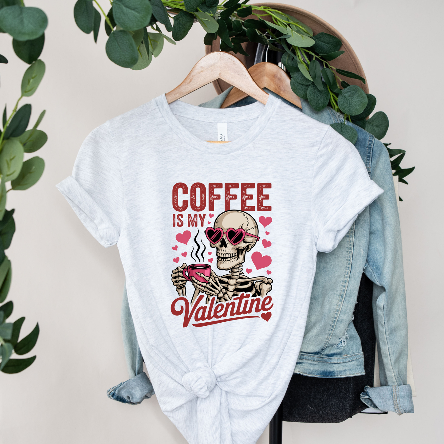 White t-shirt with skeleton design and 'Coffee is my Valentine' text on a hanger with a denim jacket and greenery. coffee shirt, coffee lover tee, valentine shirt, funny valentine shirt, coffee is my valentine, cute coffee shirt, unisex graphic tee, caffeine shirt, gift for coffee lover, funny t shirt, casual tee, valentines day shirt