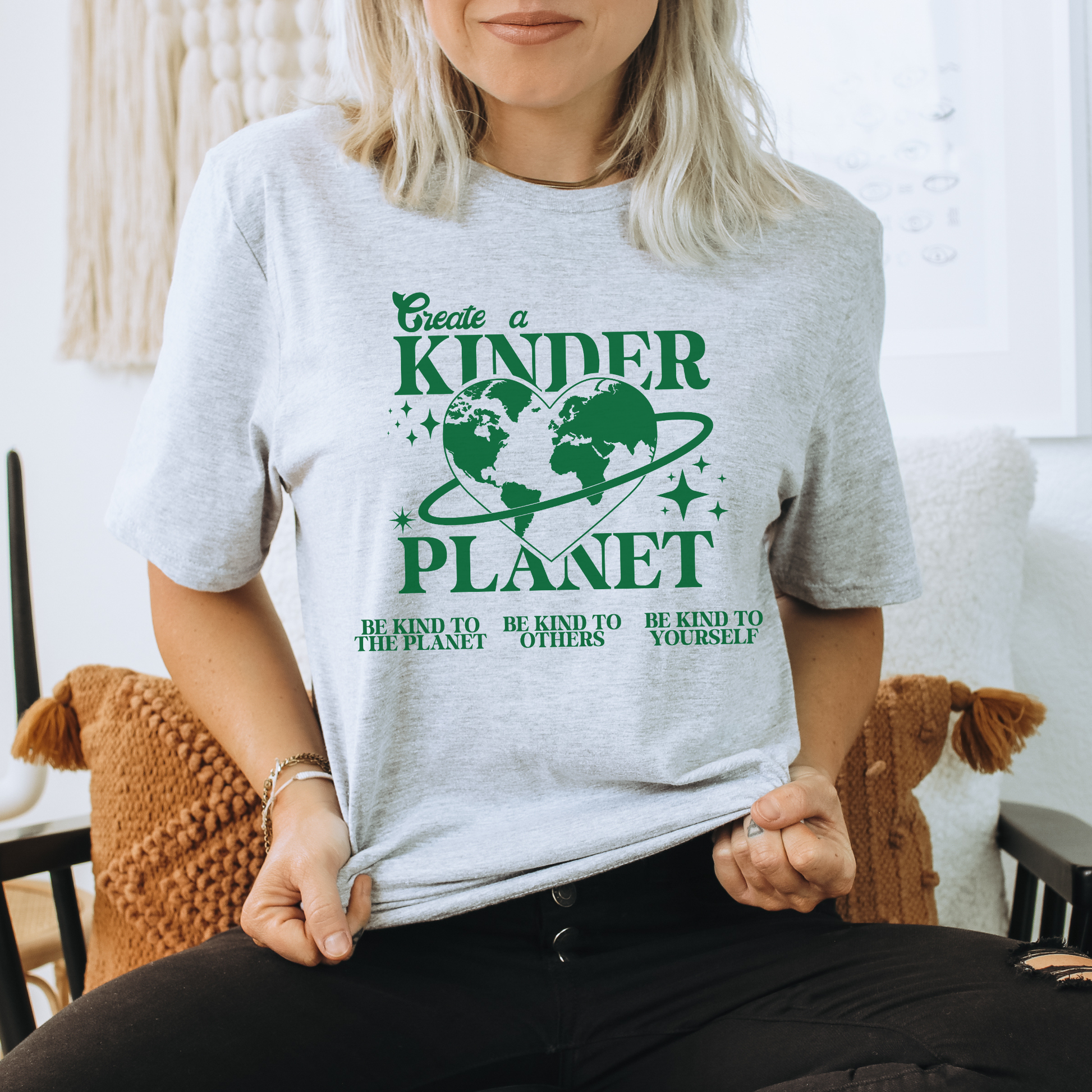 Person wearing a white t-shirt with green text and graphics, sitting in a room. Create A Kinder Planet, kinder planet shirt, kindness tee, eco awareness shirt, compassionate clothing, unisex graphic tee, environmental activism, positive change shirt, family eco tee, sustainable fashion, Indigenous brand, uplifting apparel, earth kindness, statement t-shirt, motivational tee