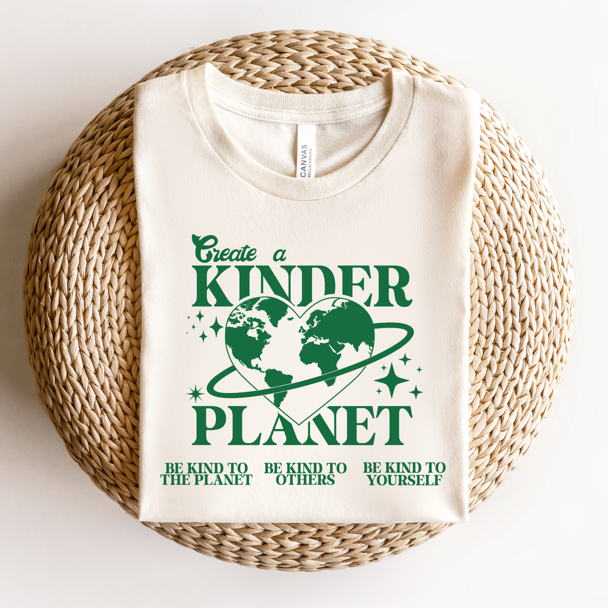 White t-shirt with green text and graphics on a woven surface. Create A Kinder Planet, kinder planet shirt, kindness tee, eco awareness shirt, compassionate clothing, unisex graphic tee, environmental activism, positive change shirt, family eco tee, sustainable fashion, Indigenous brand, uplifting apparel, earth kindness, statement t-shirt, motivational tee