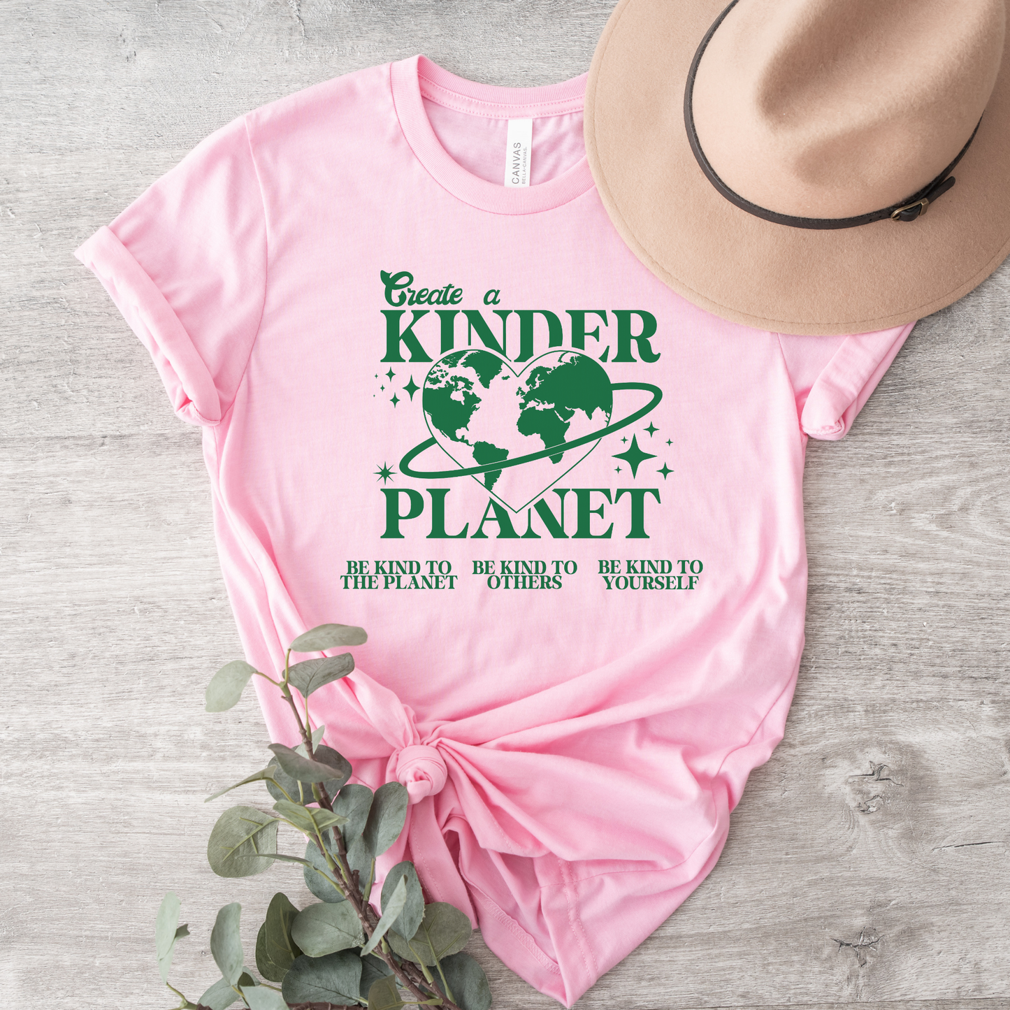 Pink t-shirt with green text and graphics on a wooden surface with a hat and plant. Create A Kinder Planet, kinder planet shirt, kindness tee, eco awareness shirt, compassionate clothing, unisex graphic tee, environmental activism, positive change shirt, family eco tee, sustainable fashion, Indigenous brand, uplifting apparel, earth kindness, statement t-shirt, motivational tee