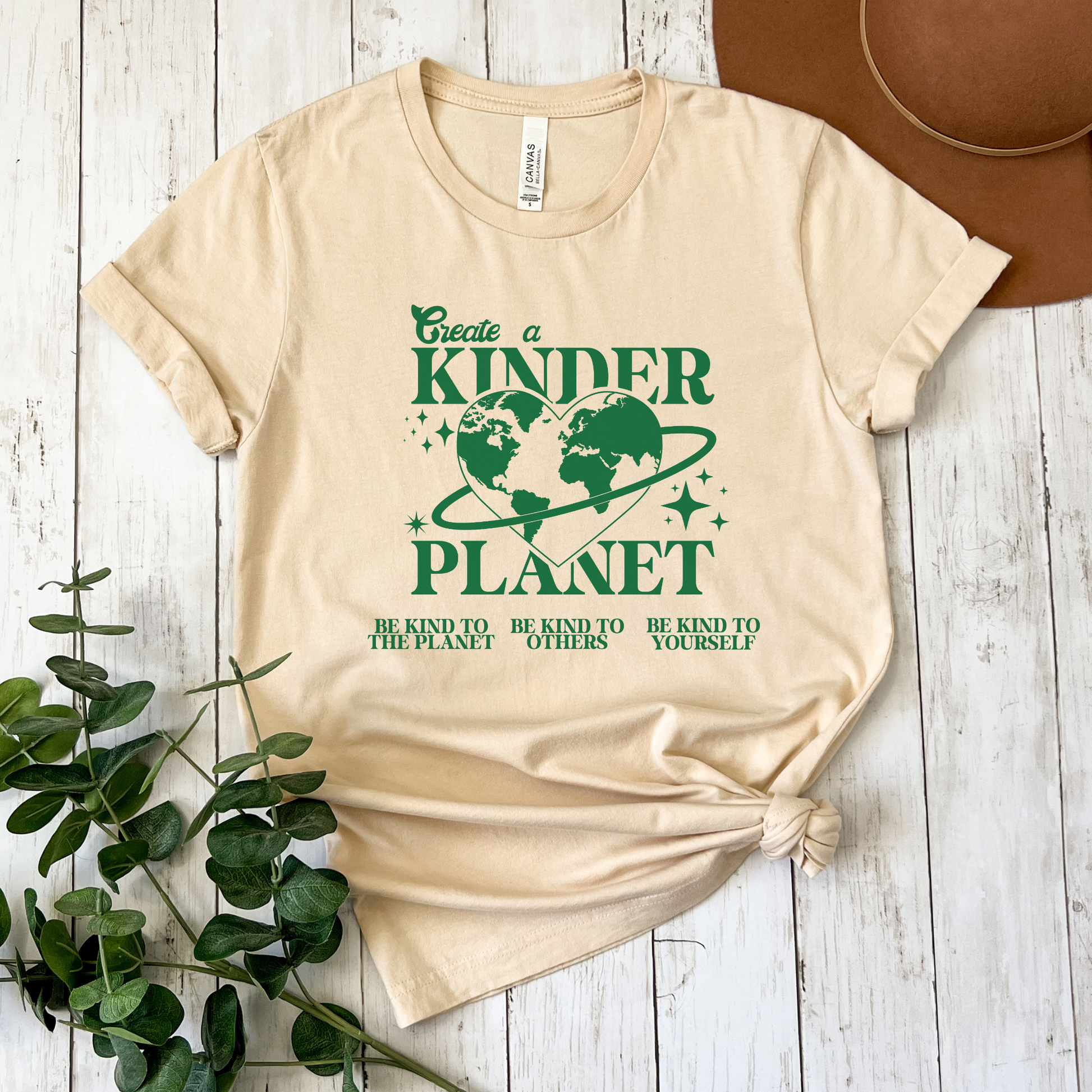 Beige t-shirt with green graphic and text on a wooden surface with a plant and hat. Create A Kinder Planet, kinder planet shirt, kindness tee, eco awareness shirt, compassionate clothing, unisex graphic tee, environmental activism, positive change shirt, family eco tee, sustainable fashion, Indigenous brand, uplifting apparel, earth kindness, statement t-shirt, motivational tee