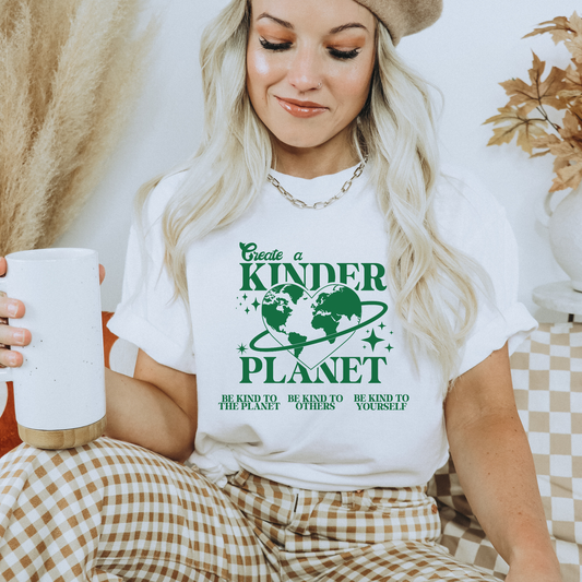 Woman wearing a white t-shirt with a green graphic and text, holding a white mug. Create A Kinder Planet, kinder planet shirt, kindness tee, eco awareness shirt, compassionate clothing, unisex graphic tee, environmental activism, positive change shirt, family eco tee, sustainable fashion, Indigenous brand, uplifting apparel, earth kindness, statement t-shirt, motivational tee