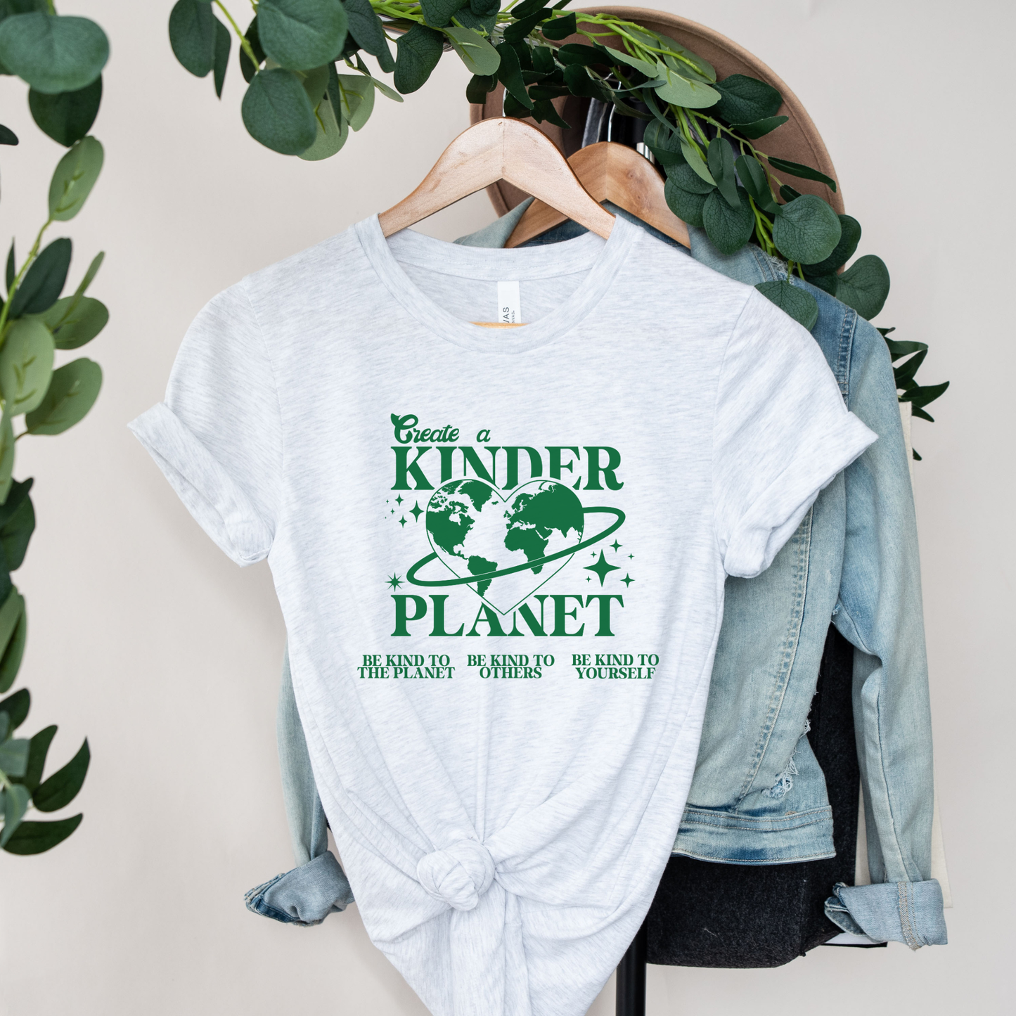 White t-shirt with green text and graphics on a hanger with a denim jacket and green leaves in the background. Create A Kinder Planet, kinder planet shirt, kindness tee, eco awareness shirt, compassionate clothing, unisex graphic tee, environmental activism, positive change shirt, family eco tee, sustainable fashion, Indigenous brand, uplifting apparel, earth kindness, statement t-shirt, motivational tee