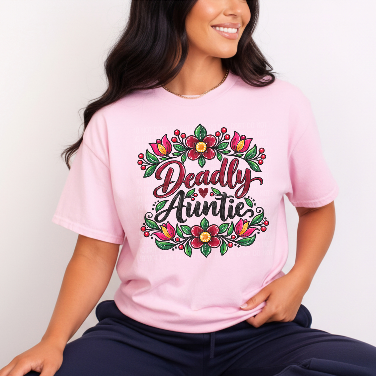A woman wearing a light pink t-shirt with a colorful graphic that reads 'Deadly Auntie' surrounded by flowers, paired with dark navy pants, set against a plain light background.
