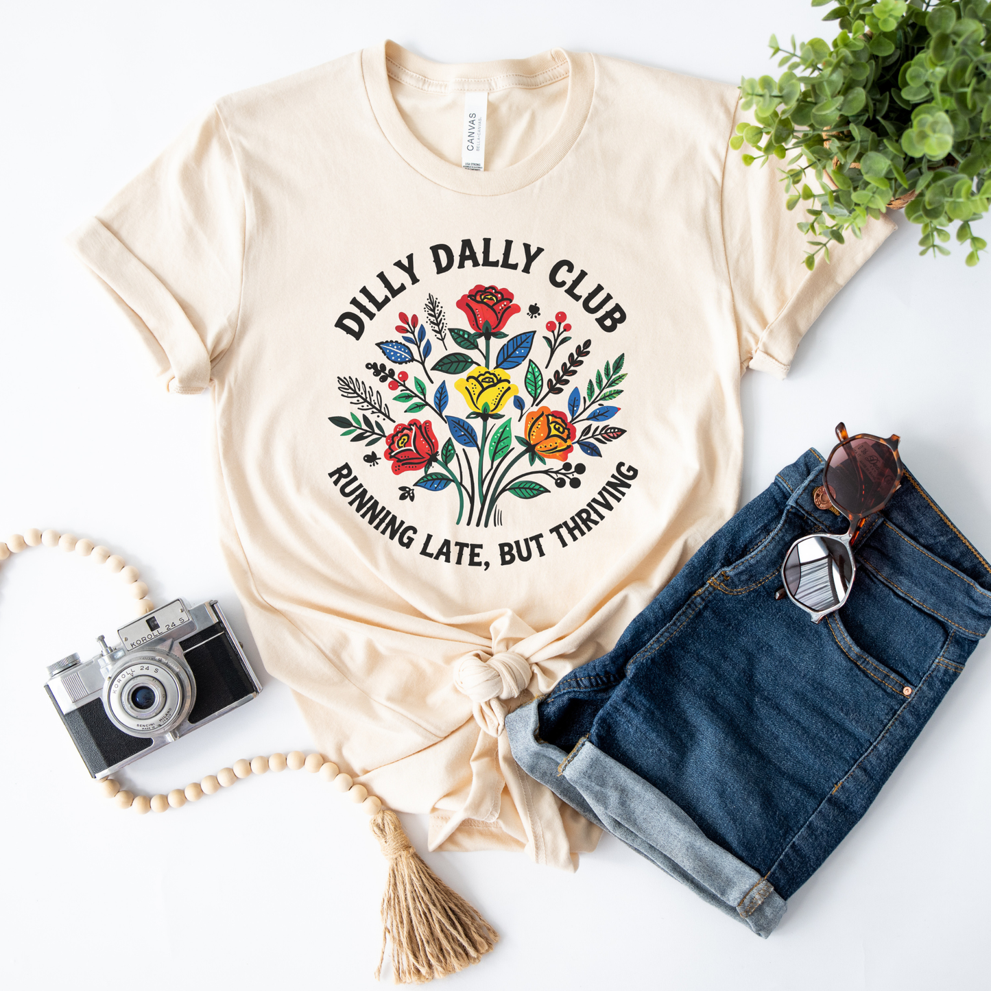 Dilly Dally Club Running Late, But Thriving shirt design featuring colorful woodland-inspired floral artwork with red, yellow, orange, blue, and green flowers arranged in a bouquet. Circular text reads 'Dilly Dally Club' and 'Running Late, But Thriving' on a transparent background. Dilly Dally Club, Running Late But Thriving, Woodland Floral, Floral Shirt, Graphic Tee, Funny Shirt, Unisex Tee, Floral Graphic Tee, Indigenous Inspired, Botanical Shirt, Cottagecore Fashion, Colorful Shirt, Gift For Her