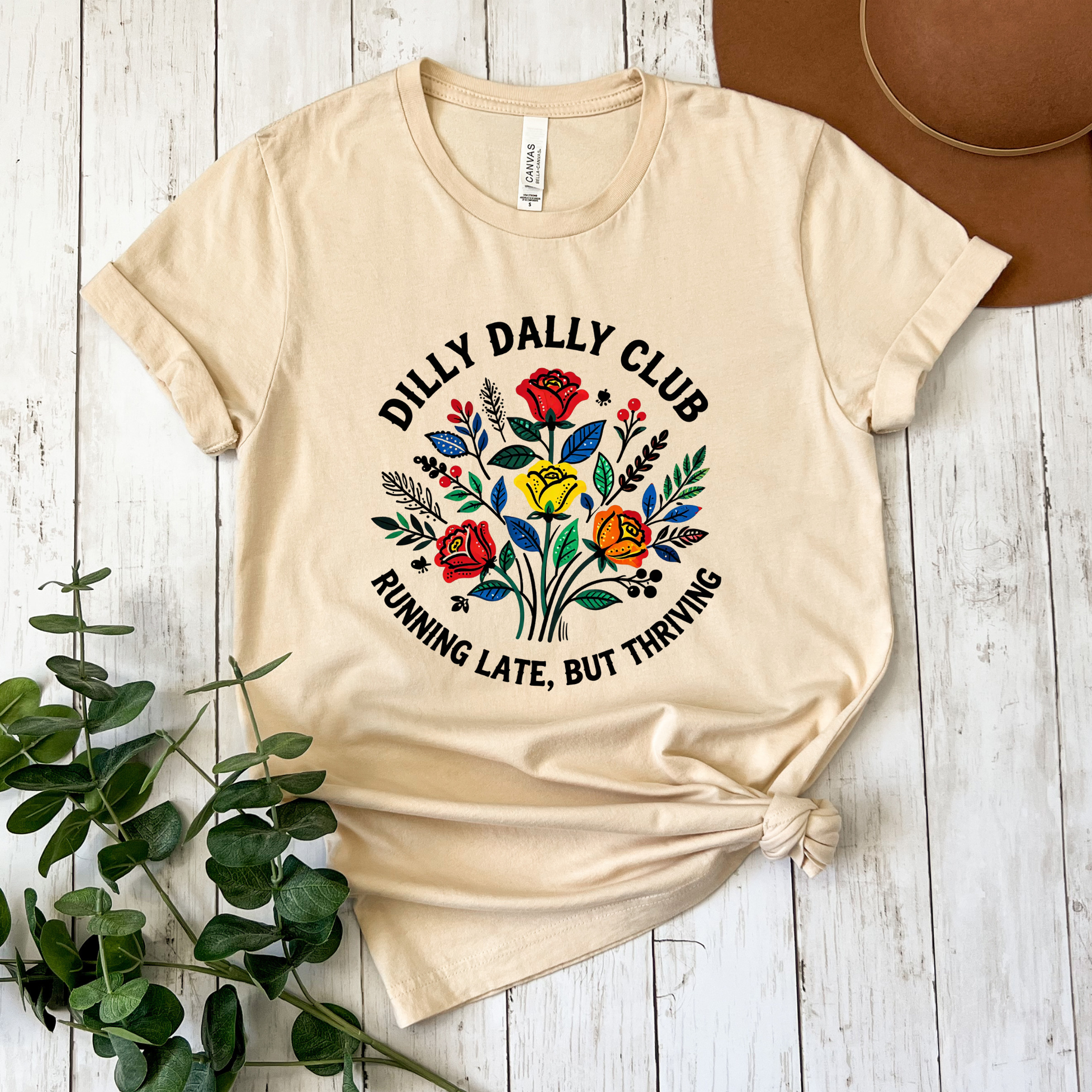 Dilly Dally Club Running Late, But Thriving shirt design featuring colorful woodland-inspired floral artwork with red, yellow, orange, blue, and green flowers arranged in a bouquet. Circular text reads 'Dilly Dally Club' and 'Running Late, But Thriving' on a transparent background. Dilly Dally Club, Running Late But Thriving, Woodland Floral, Floral Shirt, Graphic Tee, Funny Shirt, Unisex Tee, Floral Graphic Tee, Indigenous Inspired, Botanical Shirt, Cottagecore Fashion, Colorful Shirt, Gift For Her