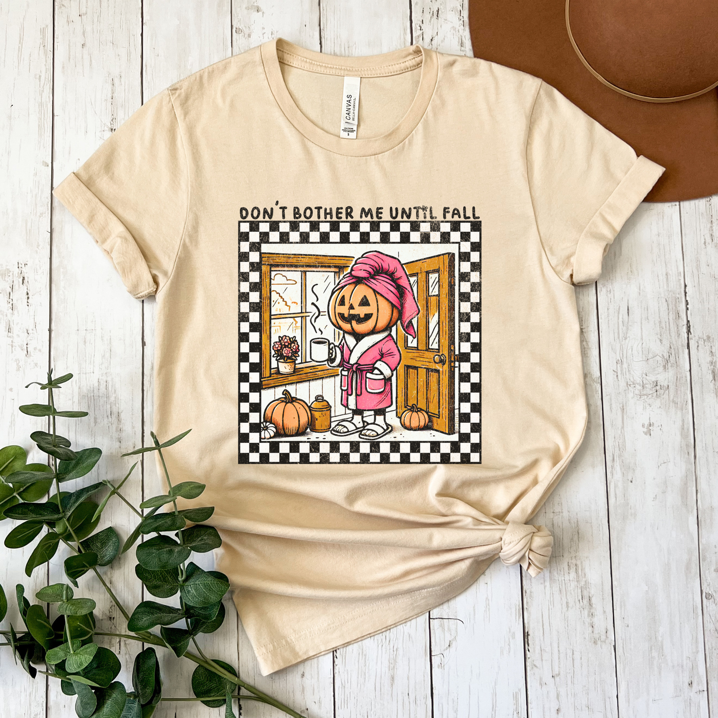 t-shirt featuring a cozy pumpkin character wearing a pink robe and towel, holding a coffee mug with the phrase "Don't Bother Me Until Fall," surrounded by autumn-inspired details and a checkered border. don't bother me until fall shirt, halloween pumpkin shirt, cozy autumn tee, fall lover t-shirt, pumpkin spice season shirt, spooky season apparel, cute pumpkin graphic tee, halloween aesthetic clothing, autumn lover gift, fall festival shirt, pumpkin patch outfit, cozy season clothing, spooky cute graphic te