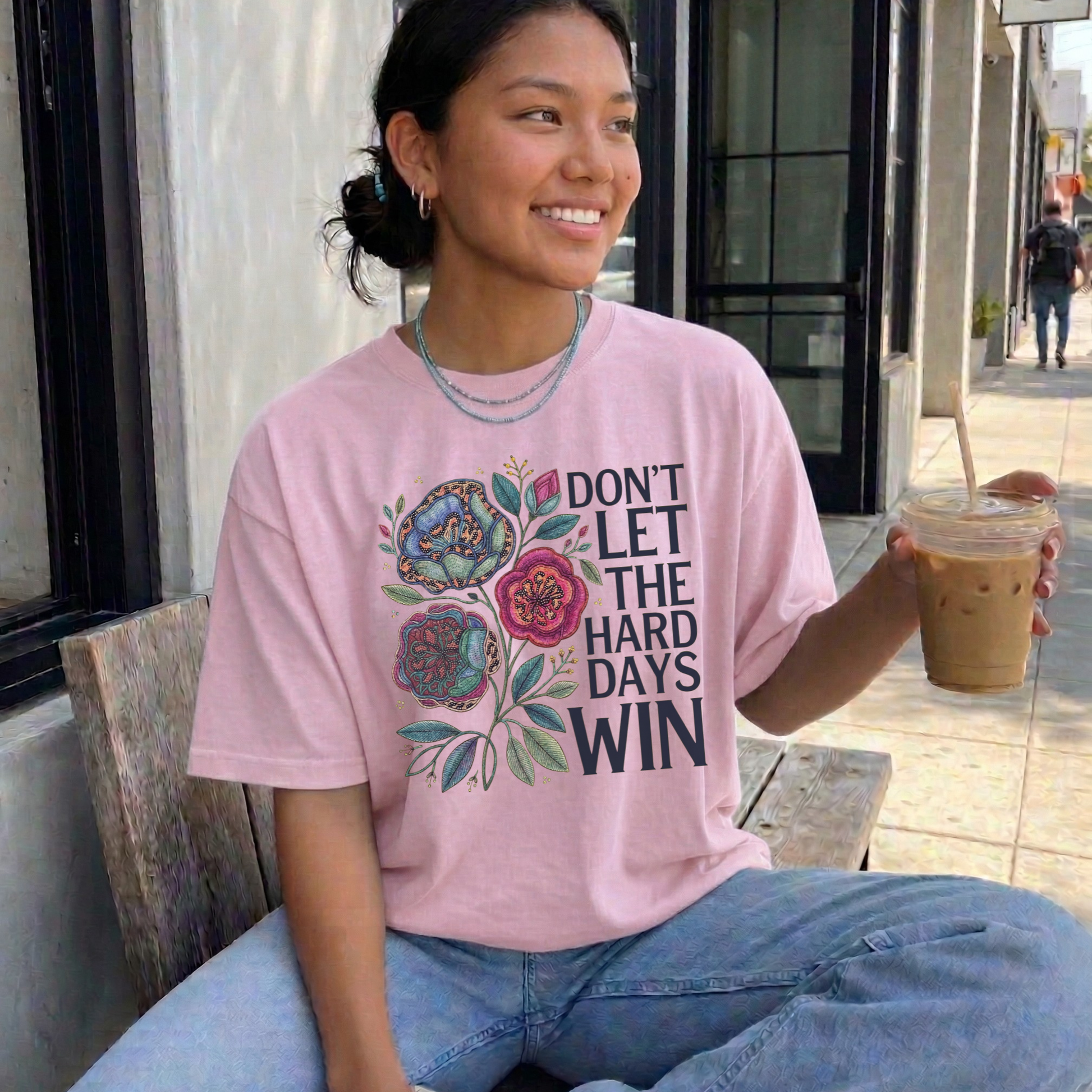 Person wearing a pink t-shirt with a motivational quote, sitting outdoors holding a drink. dont let the hard days win, inspirational shirt, motivational tee, floral graphic tee, unisex t shirt, positive quote shirt, resilience shirt, encouragement shirt, statement tee, uplifting apparel, gift idea shirt, everyday graphic tee, women’s inspirational shirt, hope shirt, feel good clothing