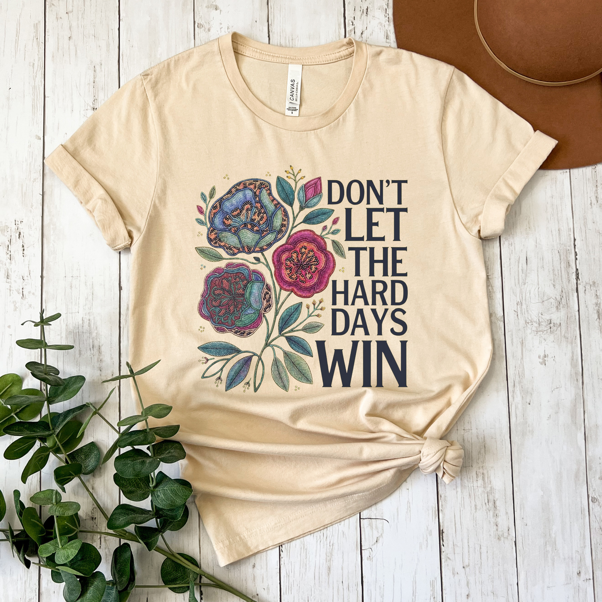 Beige t-shirt with floral design and text on a wooden surface with a plant and hat. dont let the hard days win, inspirational shirt, motivational tee, floral graphic tee, unisex t shirt, positive quote shirt, resilience shirt, encouragement shirt, statement tee, uplifting apparel, gift idea shirt, everyday graphic tee, women’s inspirational shirt, hope shirt, feel good clothing
