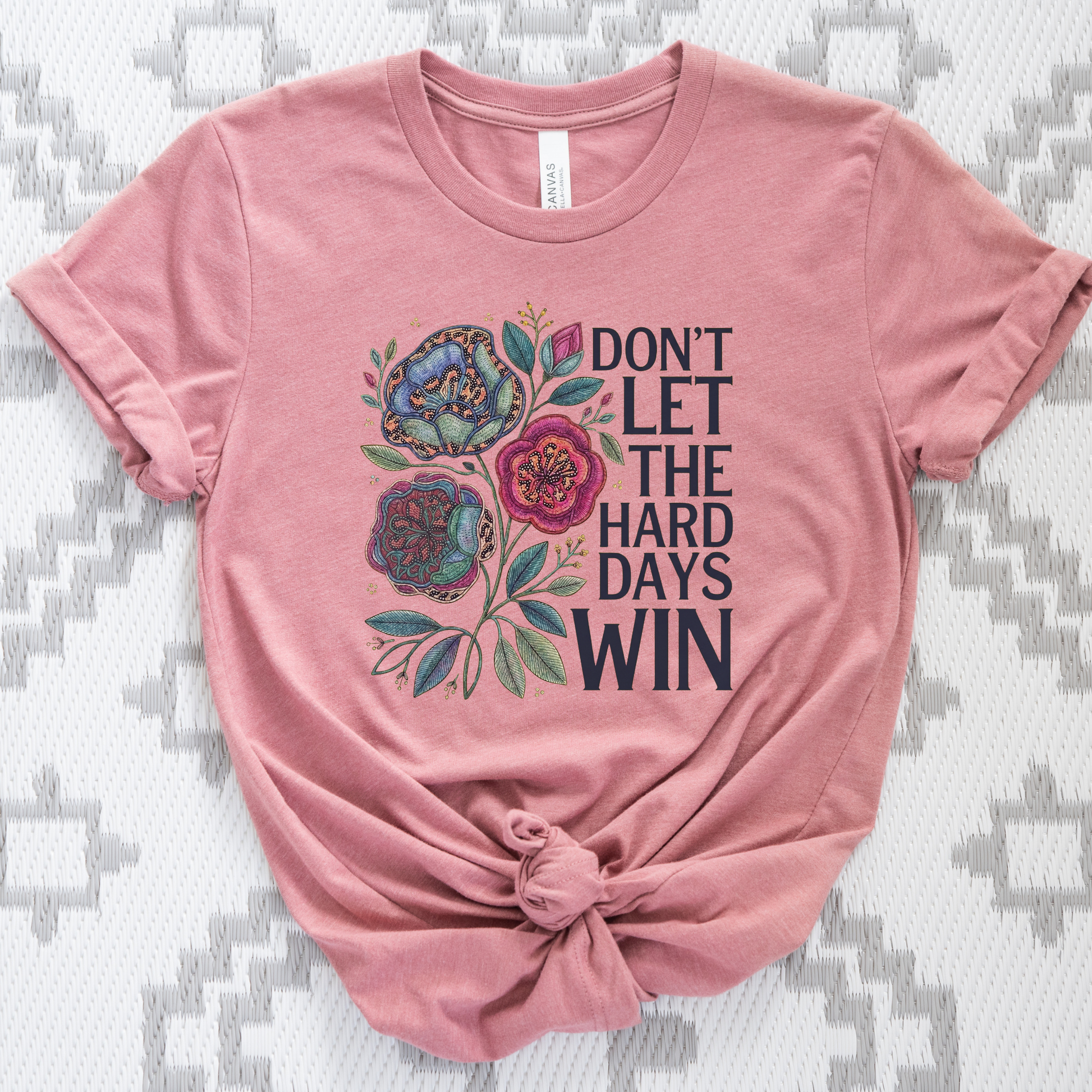 Pink t-shirt with floral design and text on a textured surface. dont let the hard days win, inspirational shirt, motivational tee, floral graphic tee, unisex t shirt, positive quote shirt, resilience shirt, encouragement shirt, statement tee, uplifting apparel, gift idea shirt, everyday graphic tee, women’s inspirational shirt, hope shirt, feel good clothing