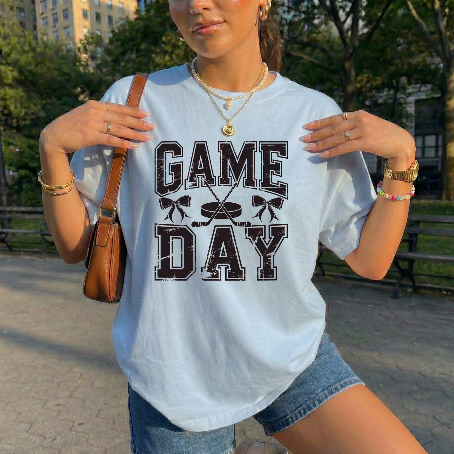A woman wearing a light blue graphic t-shirt with 'GAME DAY' printed in black varsity-style letters, posing outdoors in a park setting with denim shorts and a brown shoulder bag.
