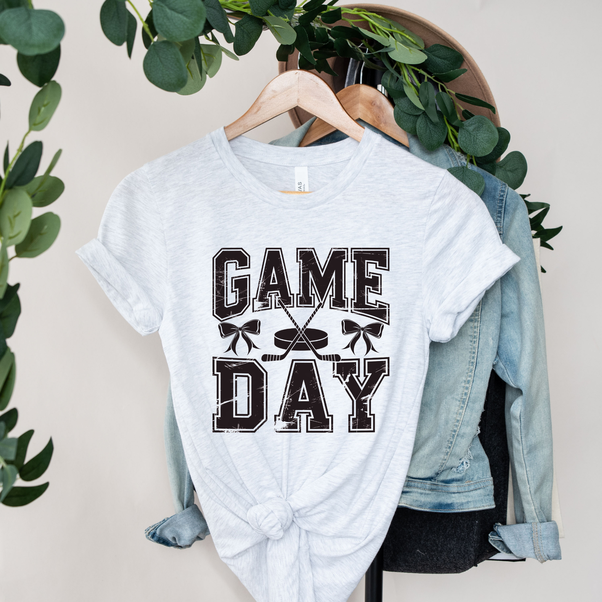 A light heather gray graphic T-shirt hanging on a wooden hanger, styled with a knotted hem and layered over a light blue denim jacket. The shirt features a distressed black graphic that reads 'GAME DAY' with crossed hockey sticks.