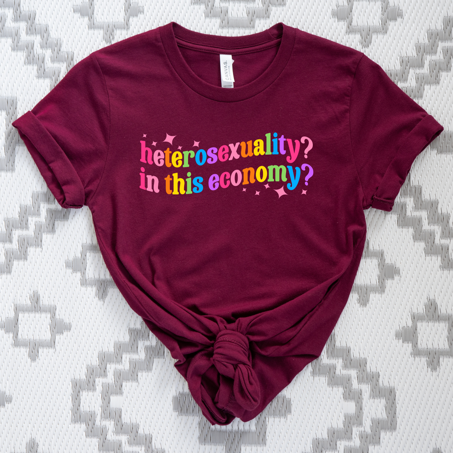 Maroon t-shirt with colorful text on a patterned background. heterosexuality in this economy, funny lgbt shirt, queer humor tee, sarcastic shirt, pride statement tee, unisex graphic tee, funny quote shirt, LGBTQ apparel, humor t shirt, statement tee, witty pride shirt, sarcastic graphic tee, queer fashion, funny unisex shirt, pride humor shirt