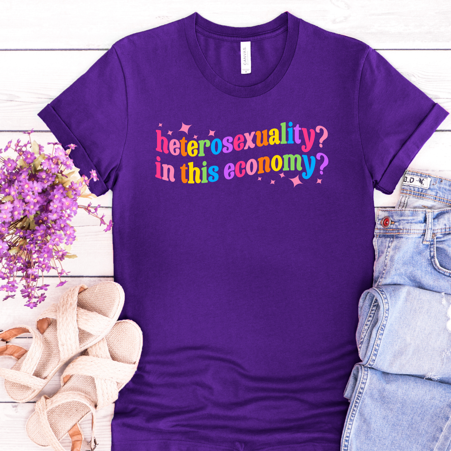 Purple t-shirt with colorful text 'heterosexuality? in this economy?' on a wooden surface with accessories.
