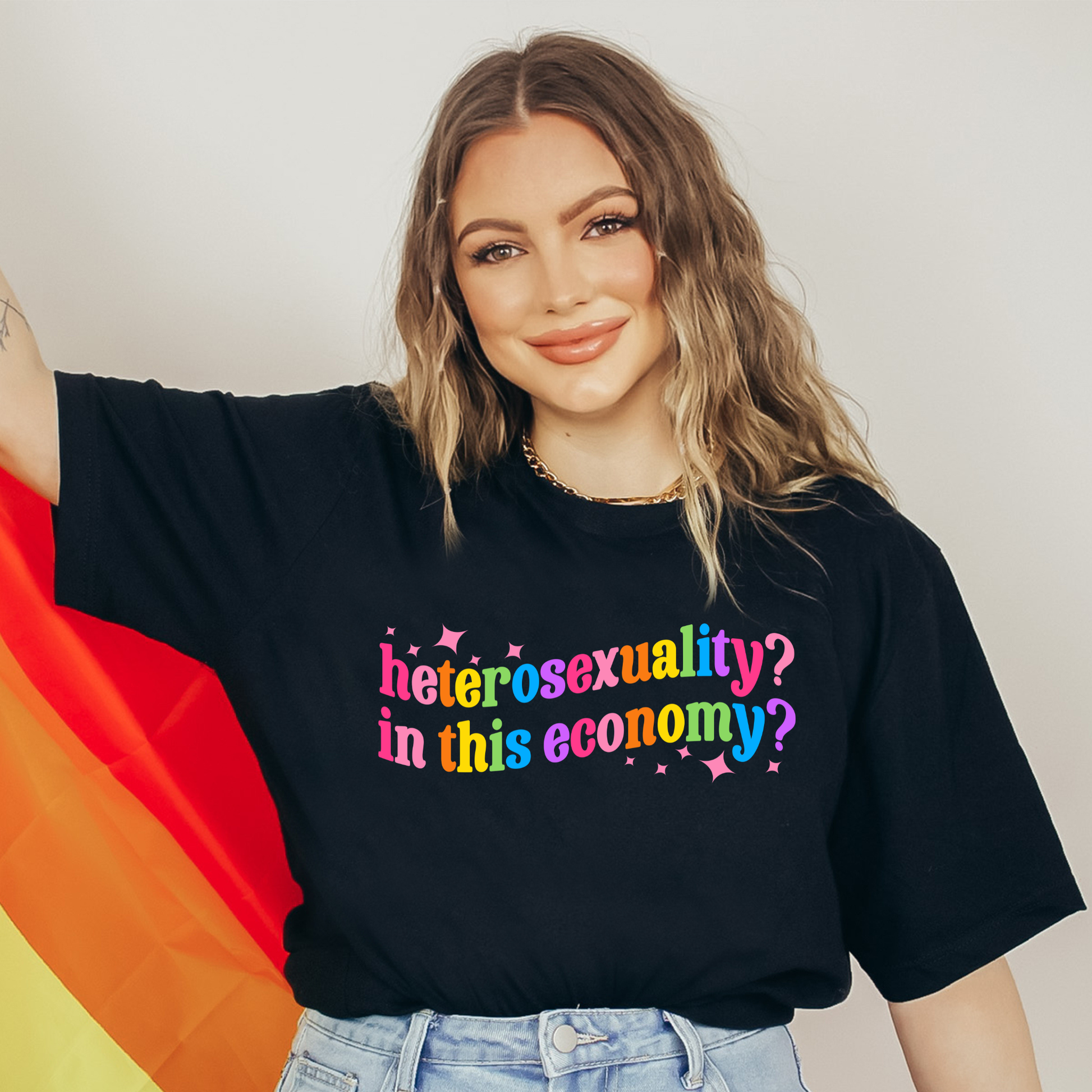 Person wearing a black t-shirt with colorful text, holding a rainbow flag. heterosexuality in this economy, funny lgbt shirt, queer humor tee, sarcastic shirt, pride statement tee, unisex graphic tee, funny quote shirt, LGBTQ apparel, humor t shirt, statement tee, witty pride shirt, sarcastic graphic tee, queer fashion, funny unisex shirt, pride humor shirt