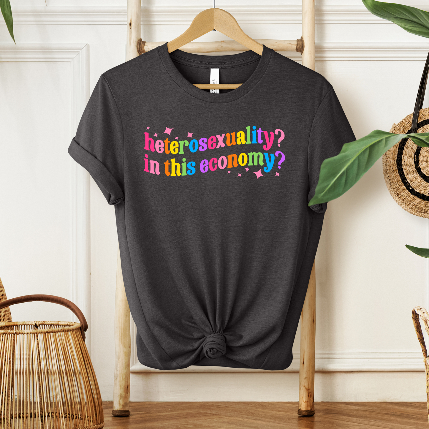 Black t-shirt with colorful text on a wooden hanger against a white wall. heterosexuality in this economy, funny lgbt shirt, queer humor tee, sarcastic shirt, pride statement tee, unisex graphic tee, funny quote shirt, LGBTQ apparel, humor t shirt, statement tee, witty pride shirt, sarcastic graphic tee, queer fashion, funny unisex shirt, pride humor shirt