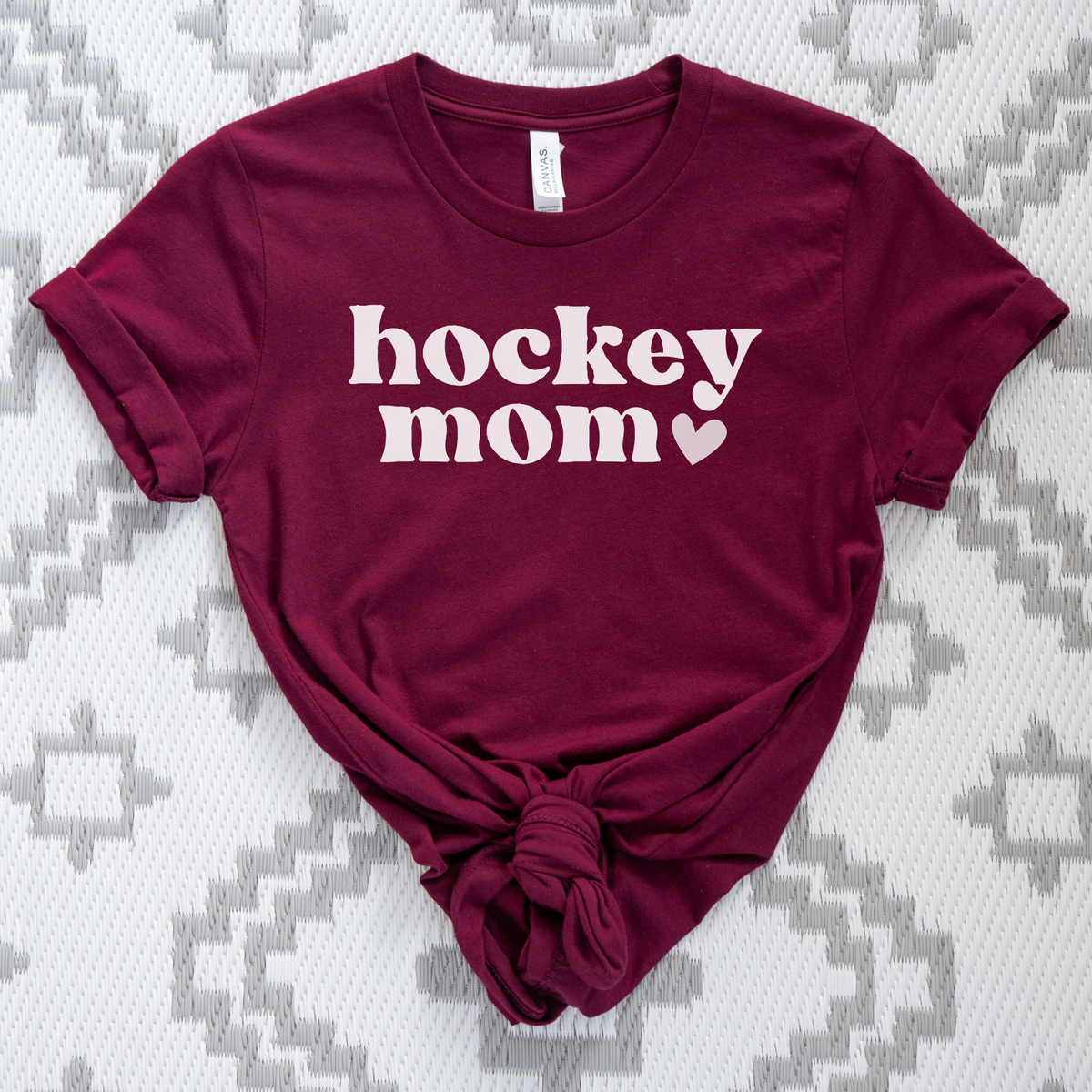 A maroon short-sleeve t-shirt with 'hockey mom' printed in white across the chest, styled with a knot at the bottom hem, laid flat on a gray and white patterned blanket.