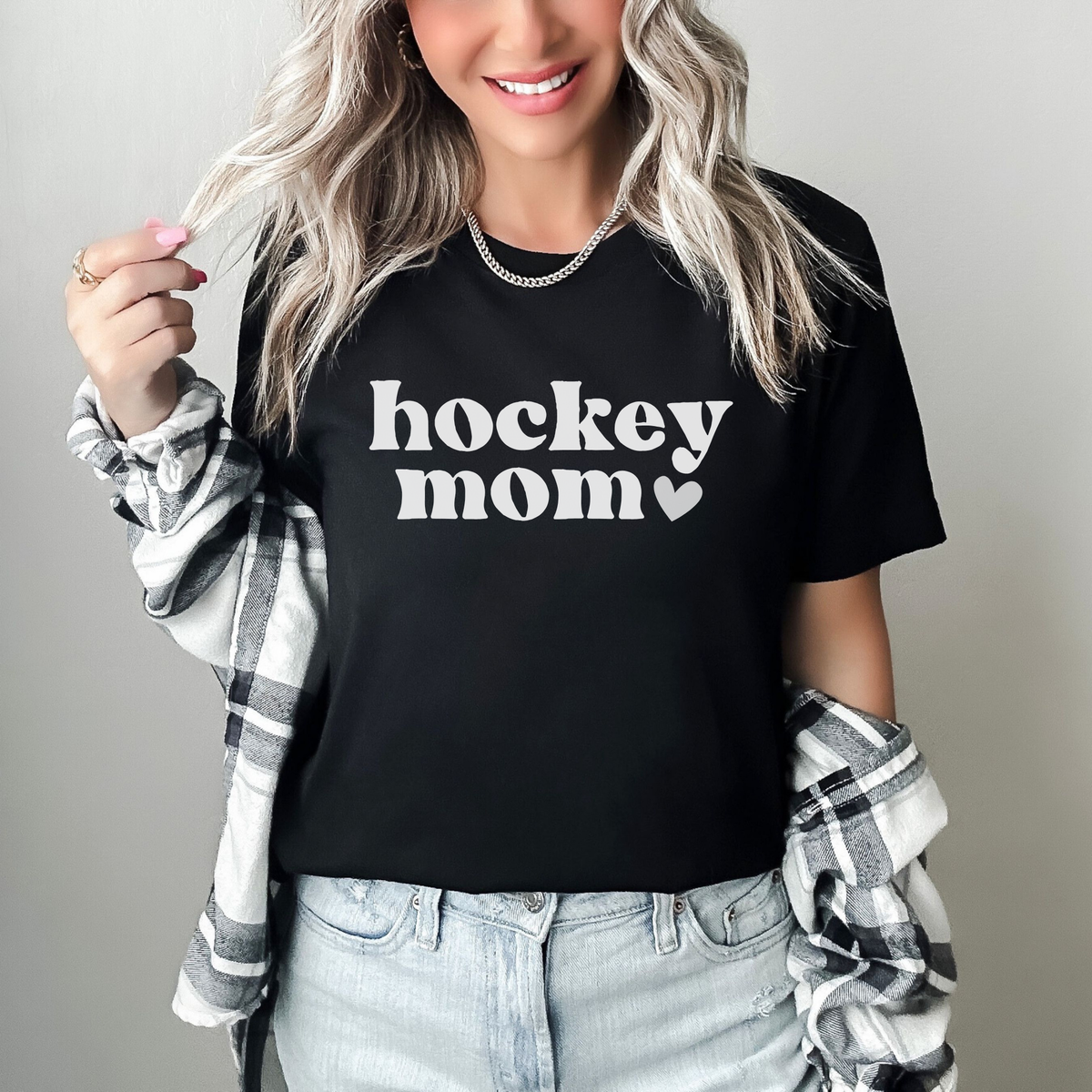 A woman wearing a black graphic t-shirt with 'hockey mom' printed in white text, styled with a black and white plaid flannel shirt and light-wash jeans against a neutral gray background. hockey mom shirt, hockey mom tee, hockey season shirt, hockey mom gift, sports mom shirt, hockey parent shirt, rink mom shirt, hockey game day, hockey tournament, hockey apparel, mom hockey shirt, hockey lover gift, retro hockey shirt