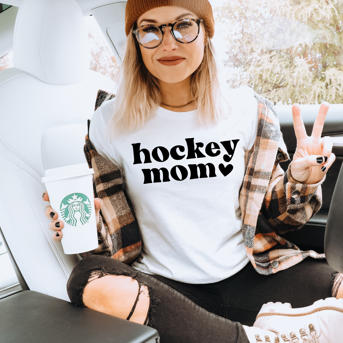 A woman sitting in a white car wearing a white graphic t-shirt that says 'hockey mom' in black text, a brown beanie, and a plaid flannel shirt. She is holding a white Starbucks coffee cup.