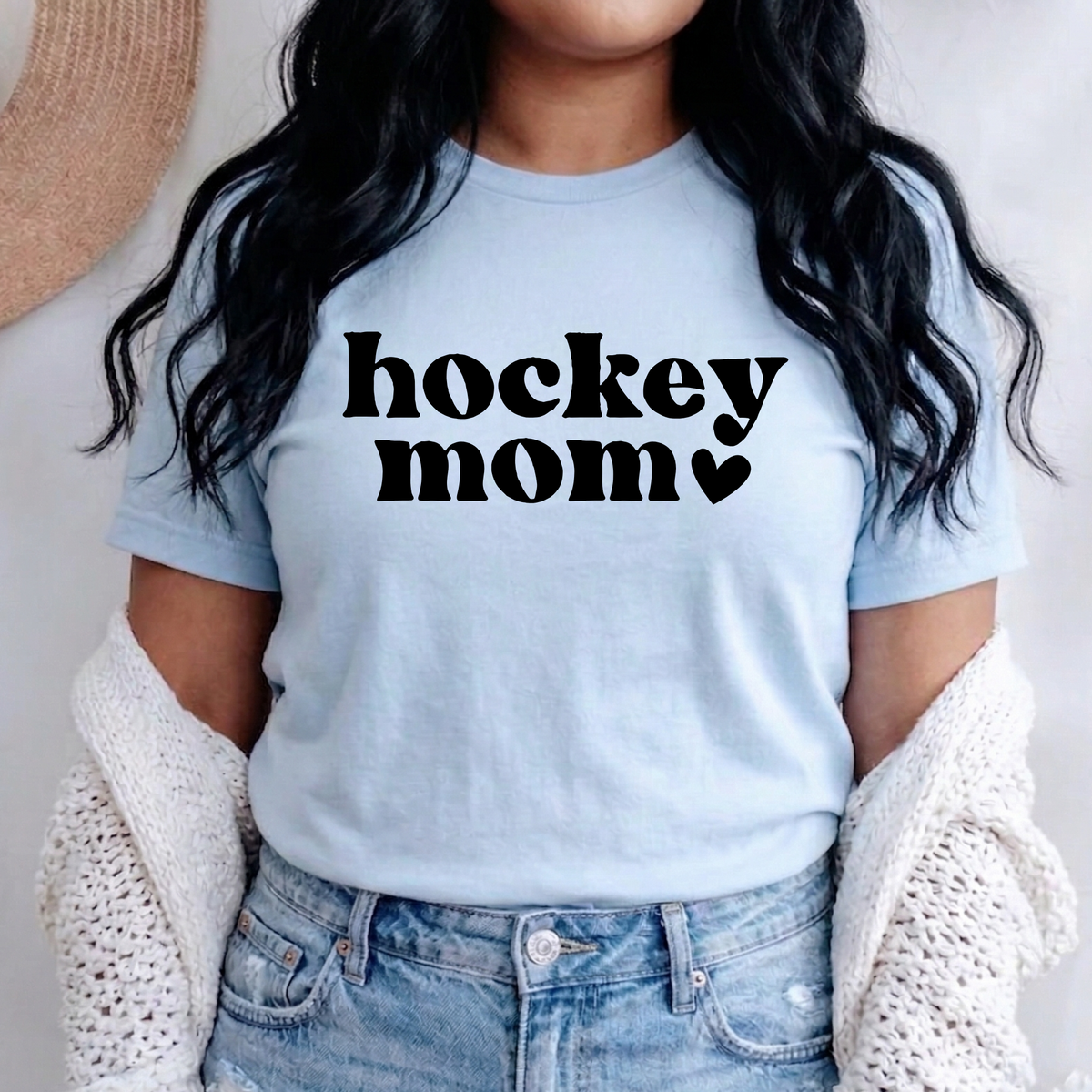A woman wearing a light blue crewneck t-shirt with 'hockey mom' printed in black text across the chest, styled with a white knit cardigan and blue jeans.