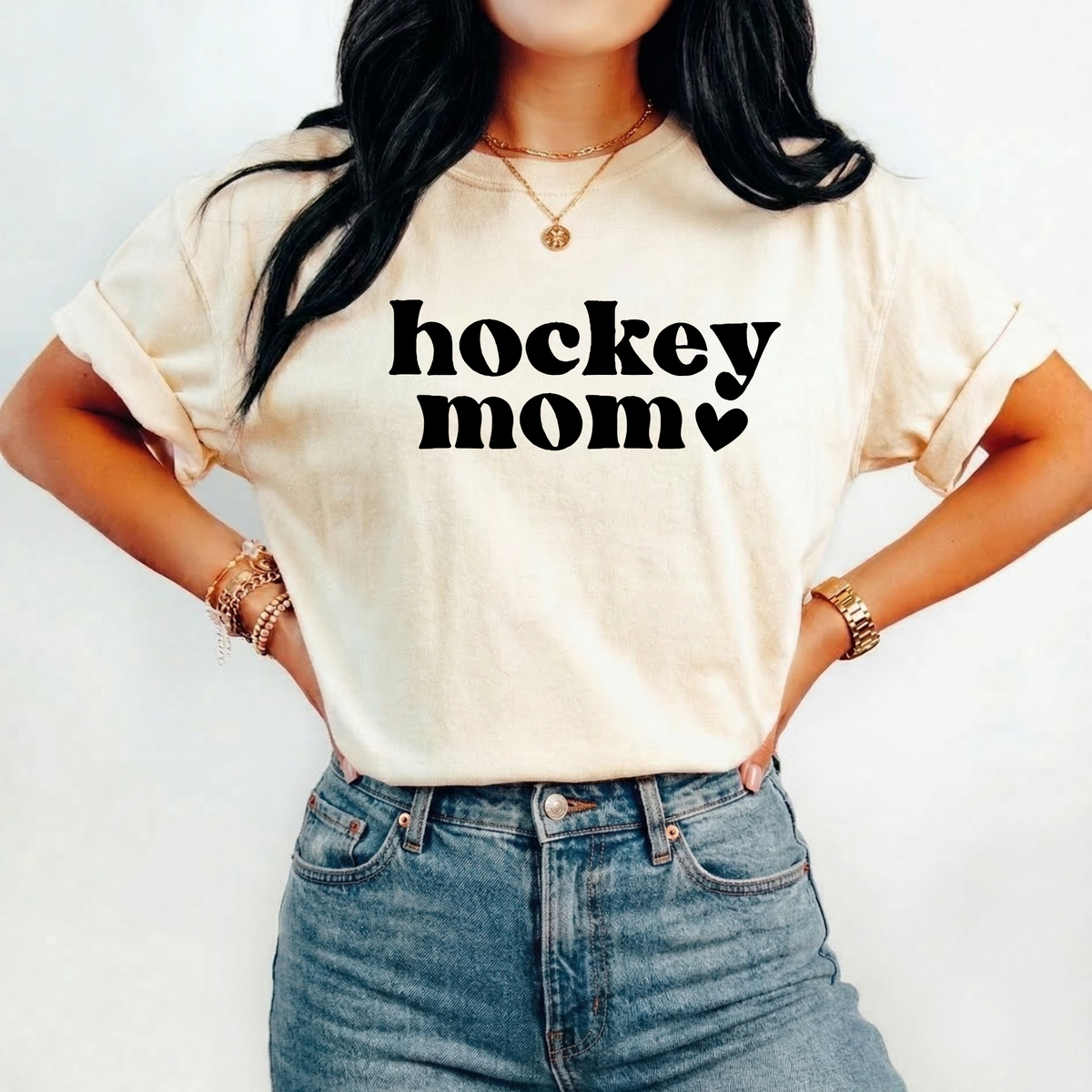 A woman wearing a cream-colored t-shirt with a black graphic that reads 'thriving' under a small leafy design, paired with blue jeans and gold jewelry, set against a plain white background. hockey mom shirt, hockey mom tee, hockey season shirt, hockey mom gift, sports mom shirt, hockey parent shirt, rink mom shirt, hockey game day, hockey tournament, hockey apparel, mom hockey shirt, hockey lover gift, retro hockey shirt