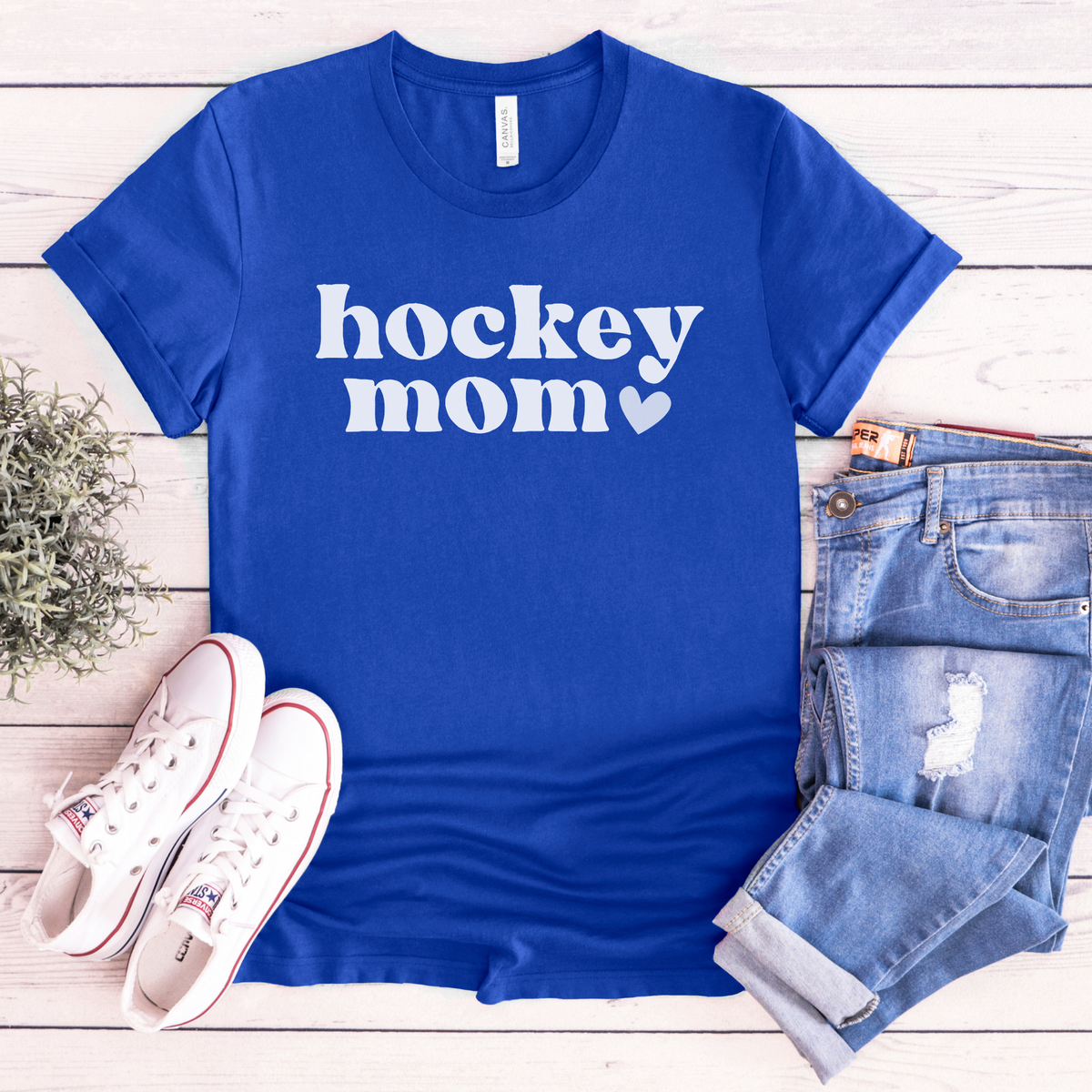 A royal blue short-sleeve T-shirt with 'hockey mom' printed in white across the chest, styled in a flat lay with light blue jeans, white sneakers, and a small plant on a whitewashed wood background.