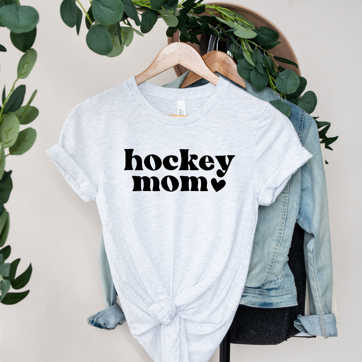 A light heather gray t-shirt with 'hockey mom' printed in black text, styled on a wooden hanger with a knotted hem, layered over a light blue denim jacket and black shorts, set against a neutral background with green foliage.